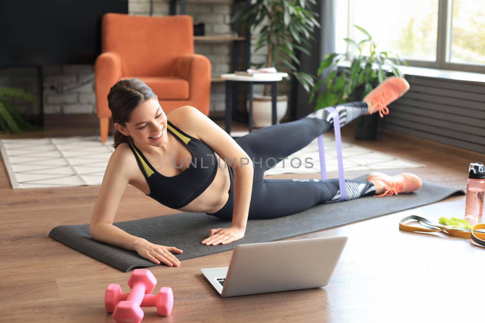Fitness beautiful slim woman doing side plank with resistance band and watching online tutorials on laptop, training in living room. Stay at home activities. by tsyhun