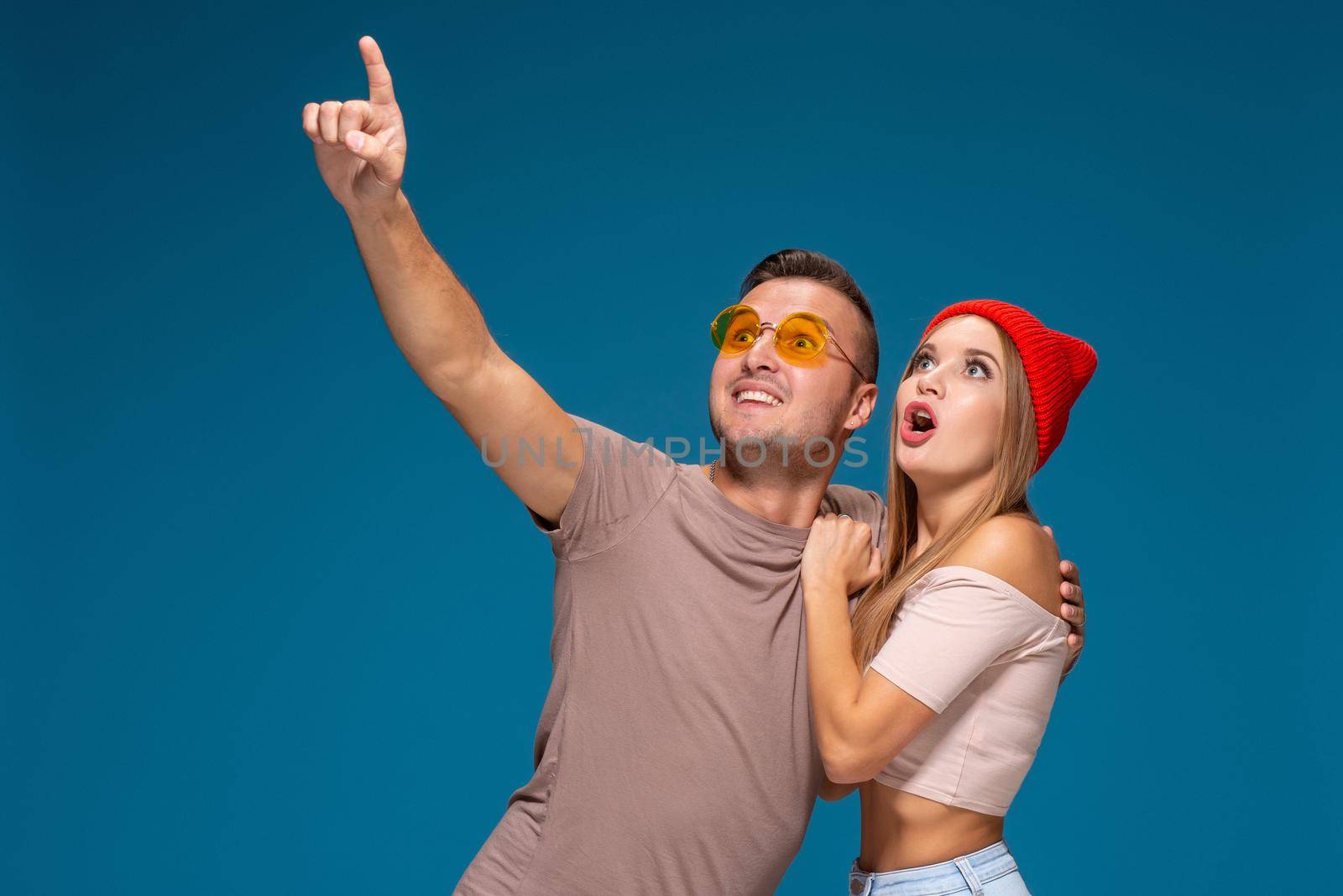 Surprised couple in casual clothes pointing with their index fingers up looking there with great astonishment isolated over blue background by nazarovsergey