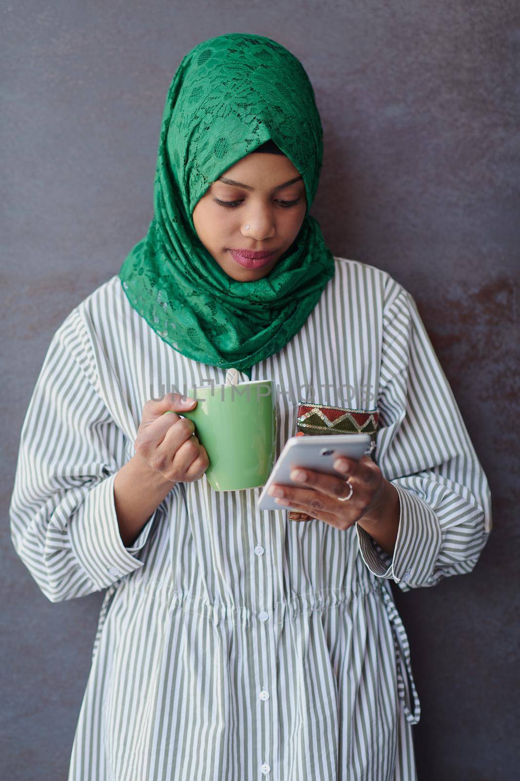 african muslim businesswoman with green hijab using mobile phone during coffee break from work in front of gray wall outside