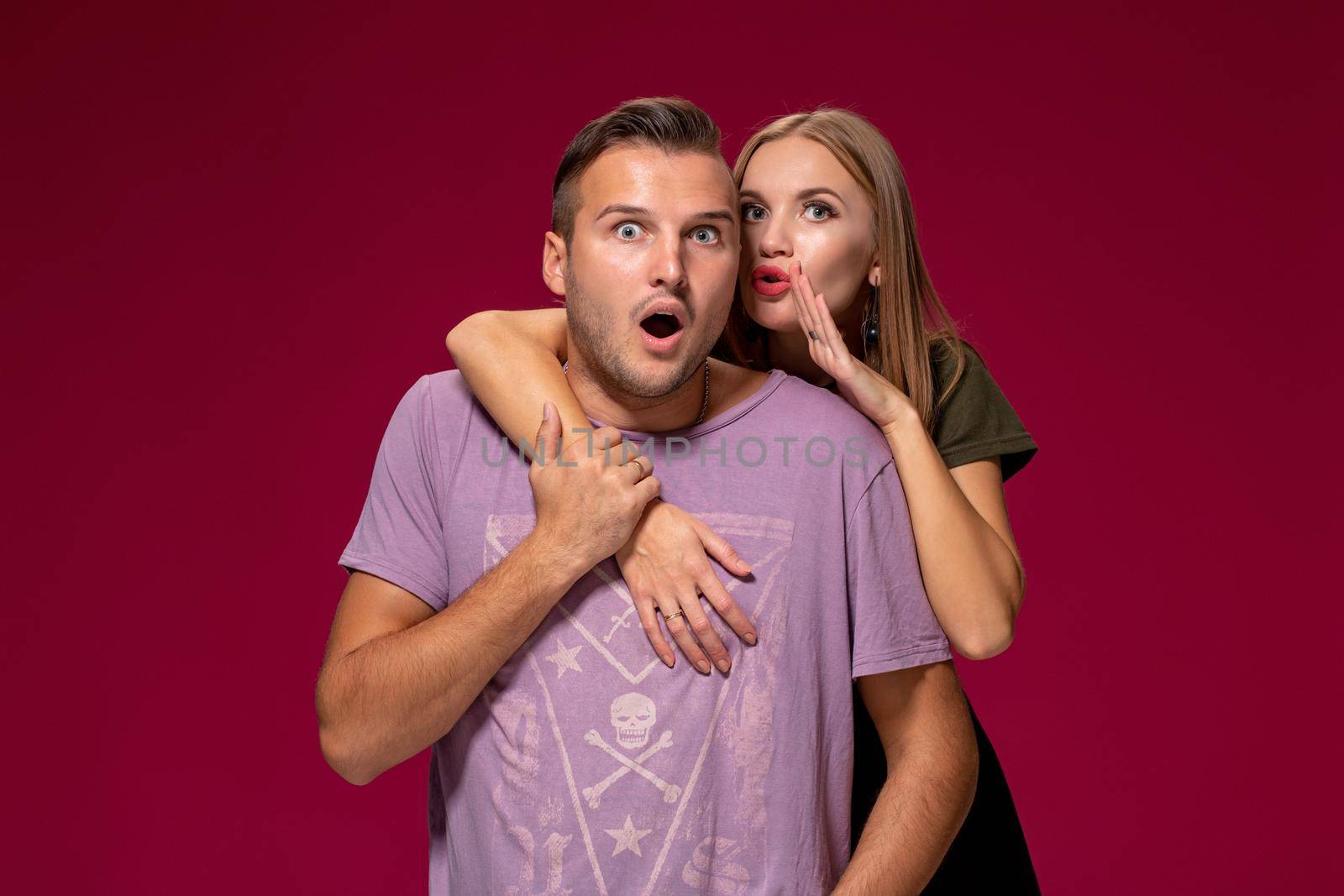 Studio shot of secret young woman and man keep index fingers on lips, stand next to each other, tell private information, ask not spread rumors and be quiet, isolated over burgundy background.