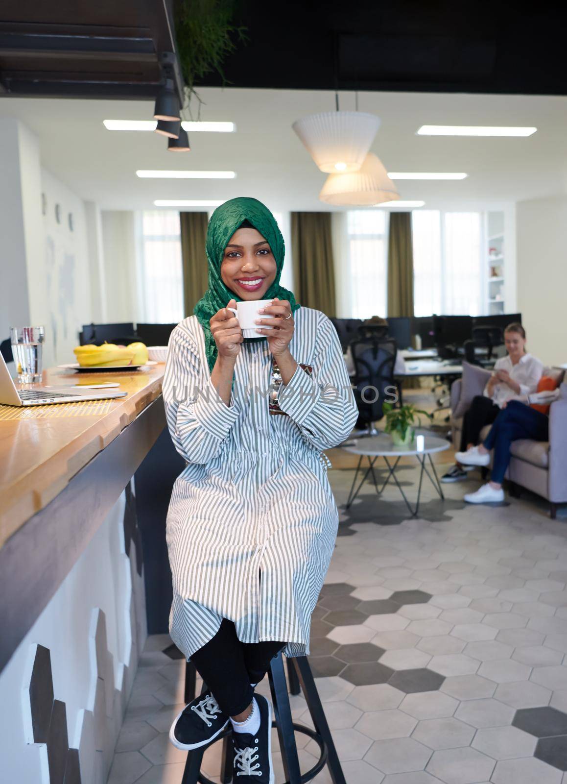 african muslim business woman wearing a green hijab drinking tea while working on laptop computer in relaxation area at modern open plan startup office. Diversity, multiracial concept