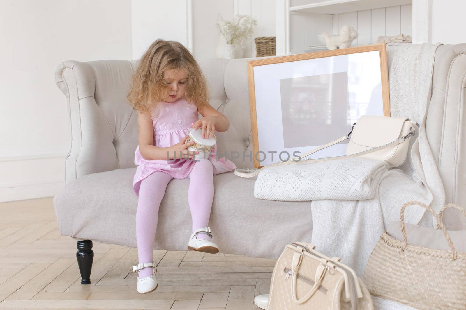 Adorable girl in pink dress trying to open box while sitting on comfortable couch near stylish bags and blank frame in elegant room