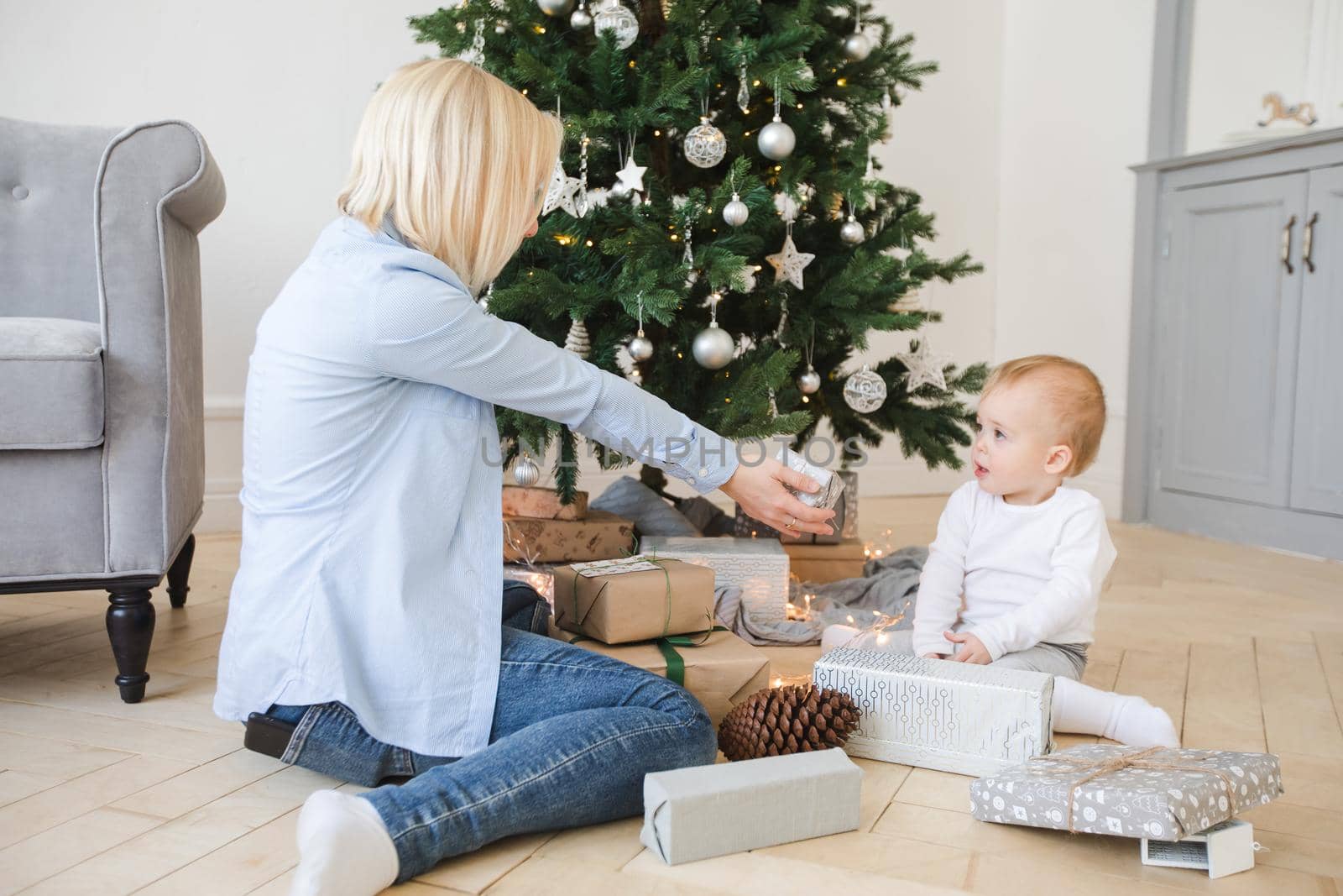 Side view of young blond woman sitting with little child on parquet near Christmas decorated tree among wrapped gifts