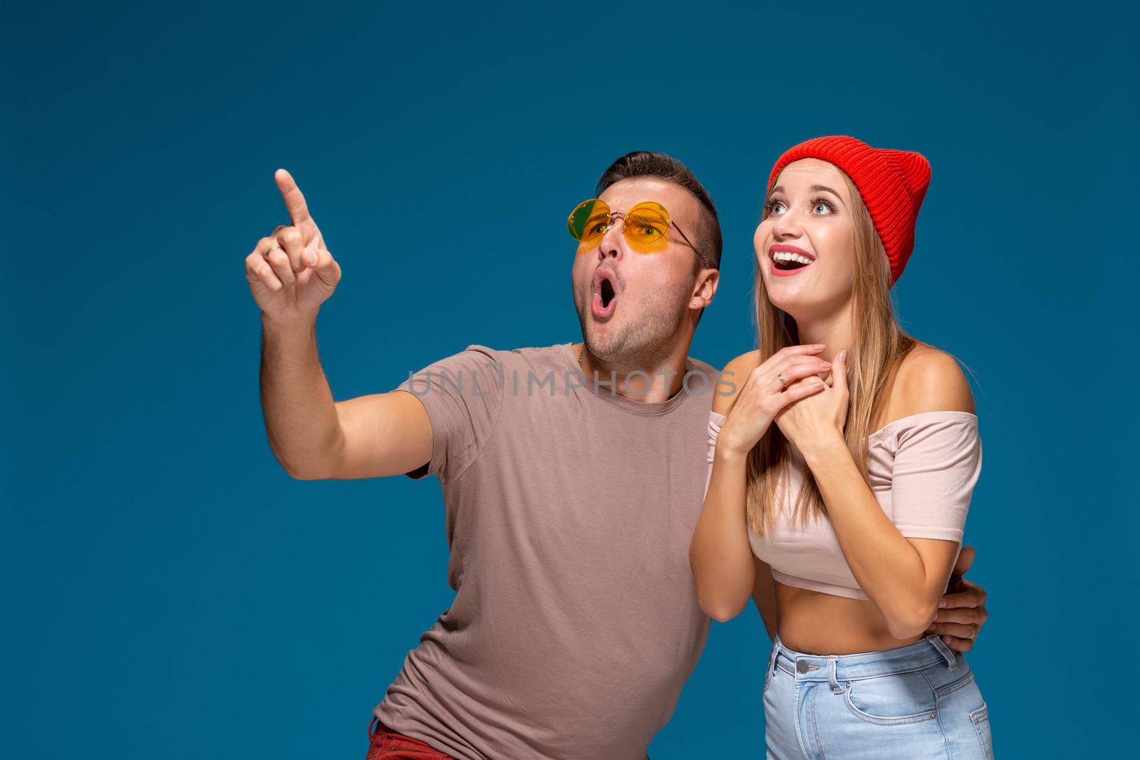 Surprised couple in casual clothes pointing with their index fingers up looking there with great astonishment isolated over blue background with copy space for your advertisement or written text.