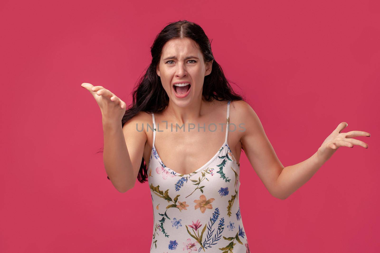 Portrait of a woman in a white dress with floral print standing on a pink wall background in studio. She swears at someone and screams loudly. People sincere emotions, lifestyle concept. Mockup copy space.
