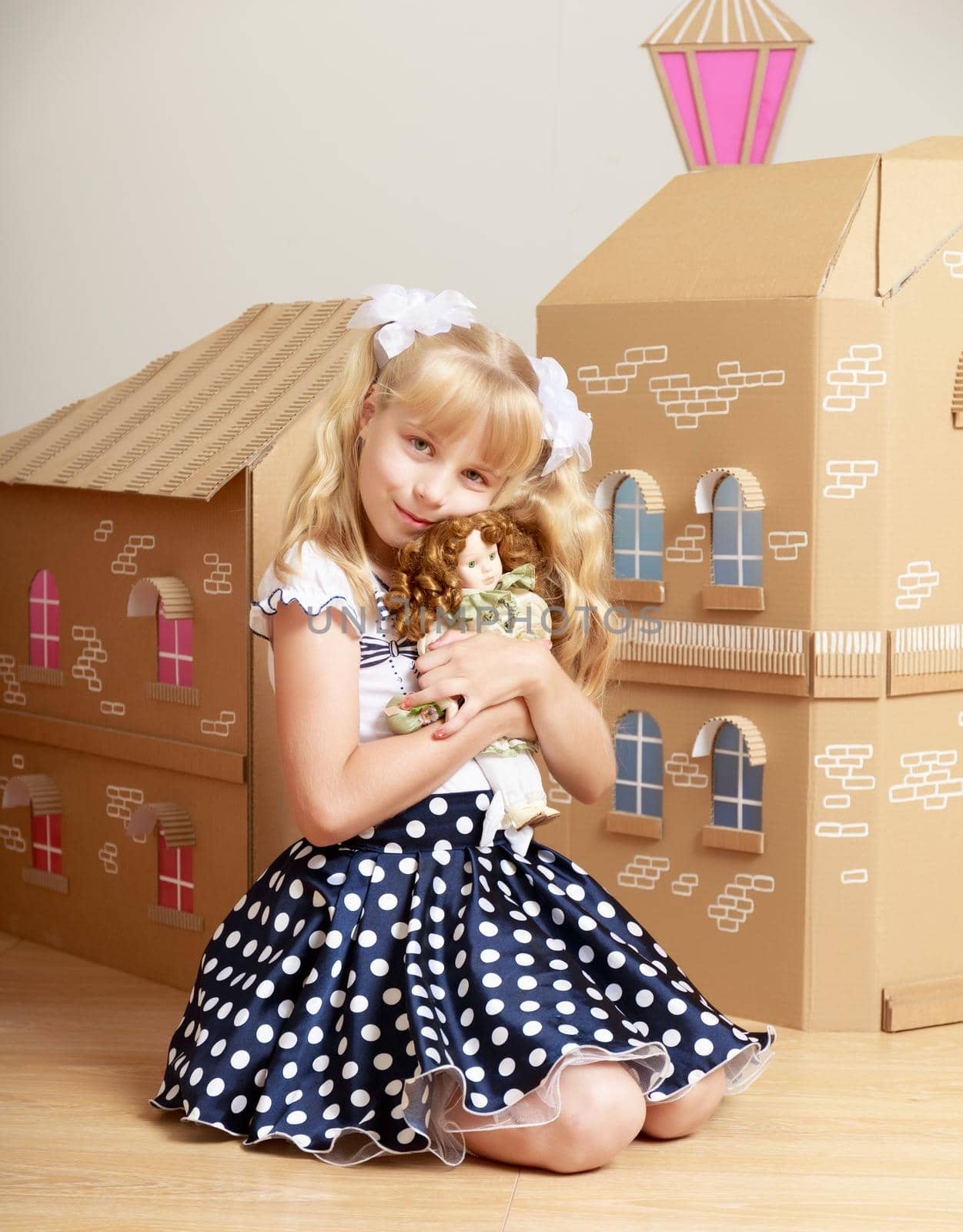 Beautiful little girl with long blonde ponytails on her head , playing with a doll near a cardboard house