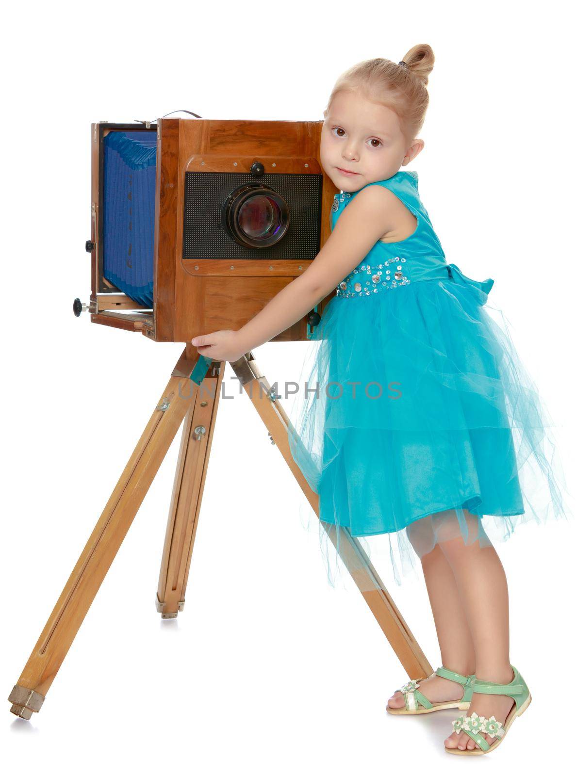 Cute little girl in blue elegant dress posing near antique wooden camera, wooden legs.Isolated on white background