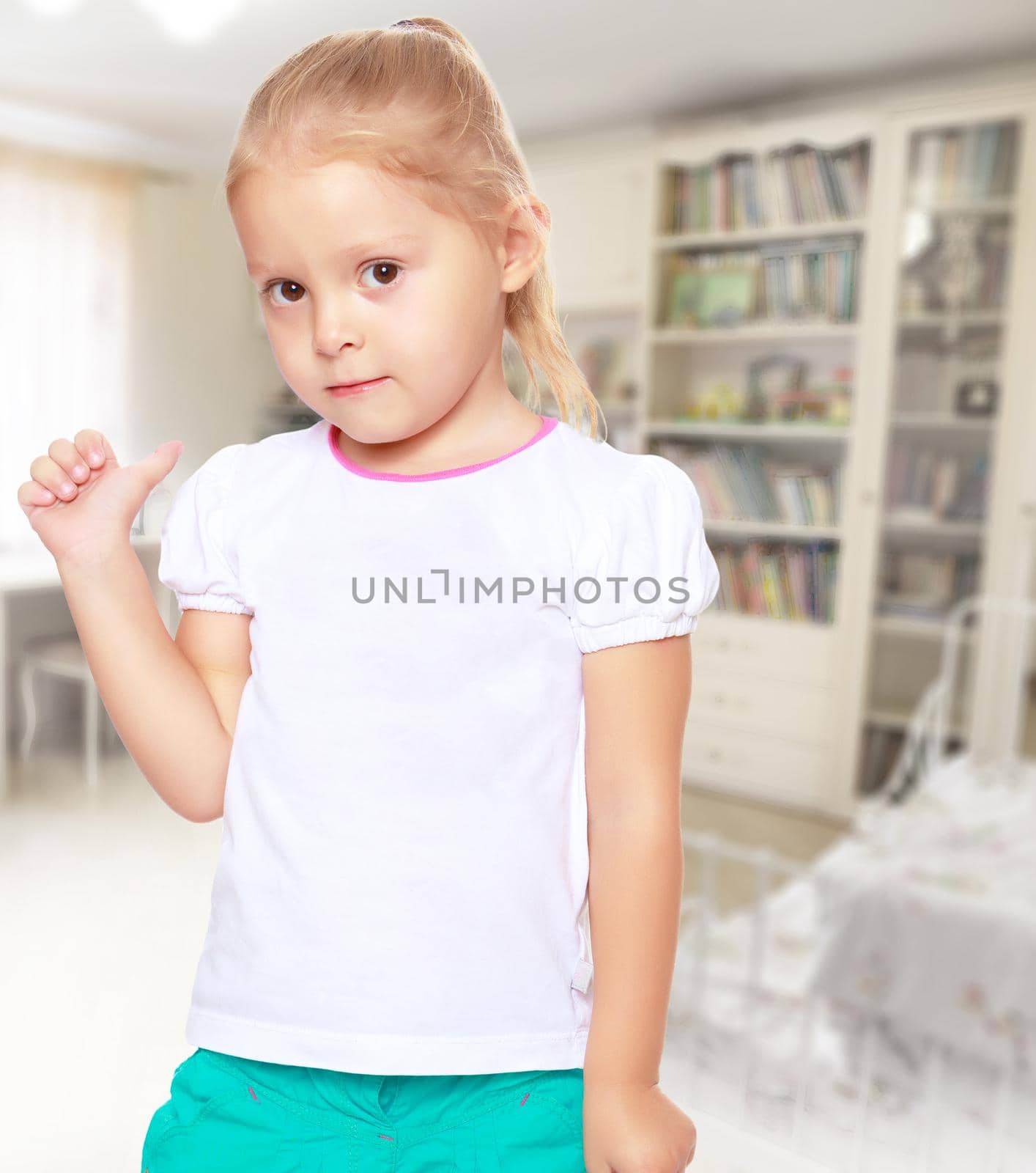 Cute little blond girl in white t-shirt . Girl shows thumb ago. In the background children's room and bookcases . The girl is preparing for school.