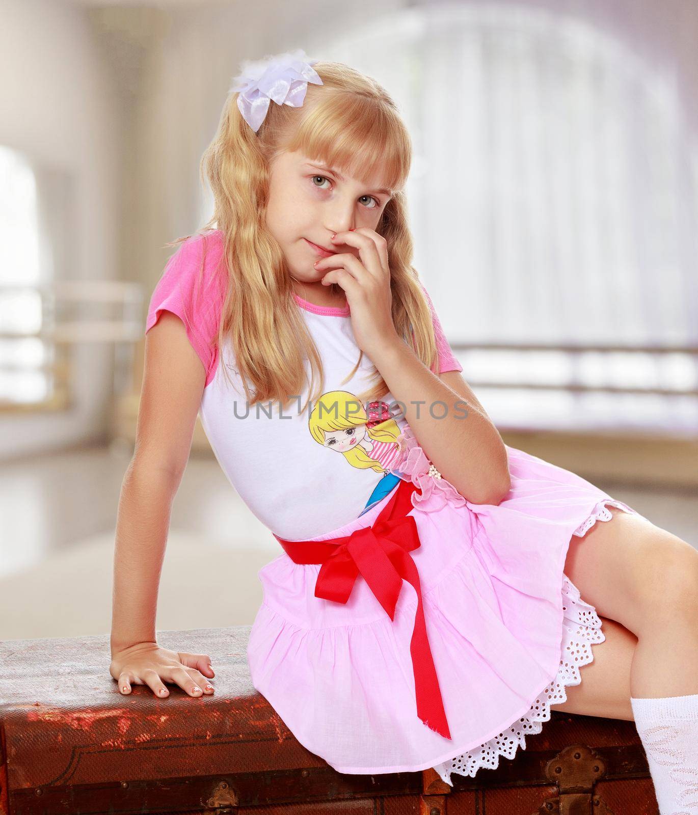 Beautiful little girl in a pink short skirt and white socks sitting on the old road suitcase.In a room with a large semi-circular window.