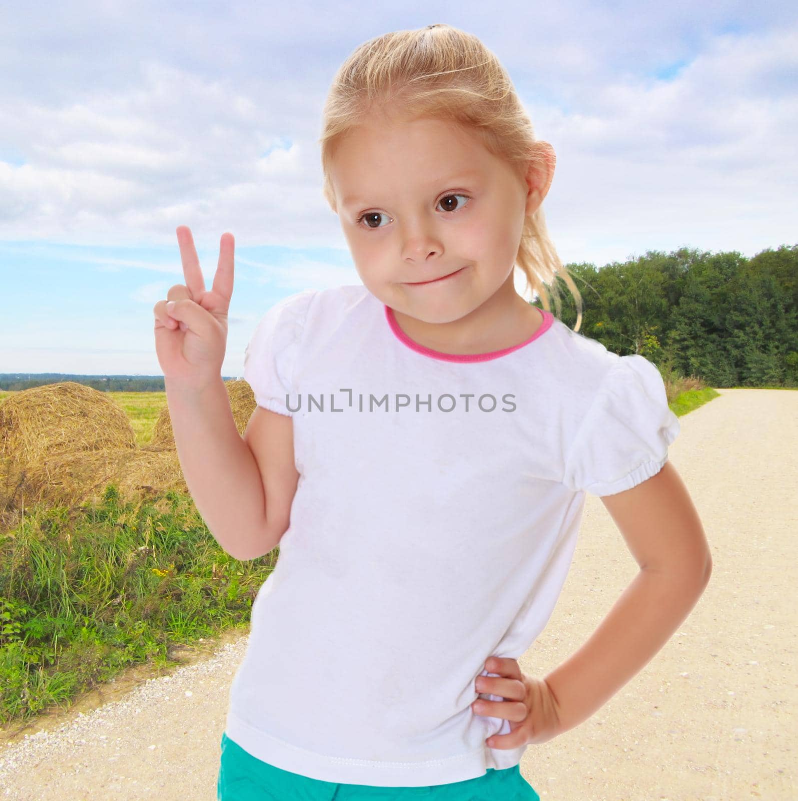 Cute little blond girl in white t-shirt . Girl shows hand gesture Victoria. In the background stretching into the distance on the road and the mown hay.