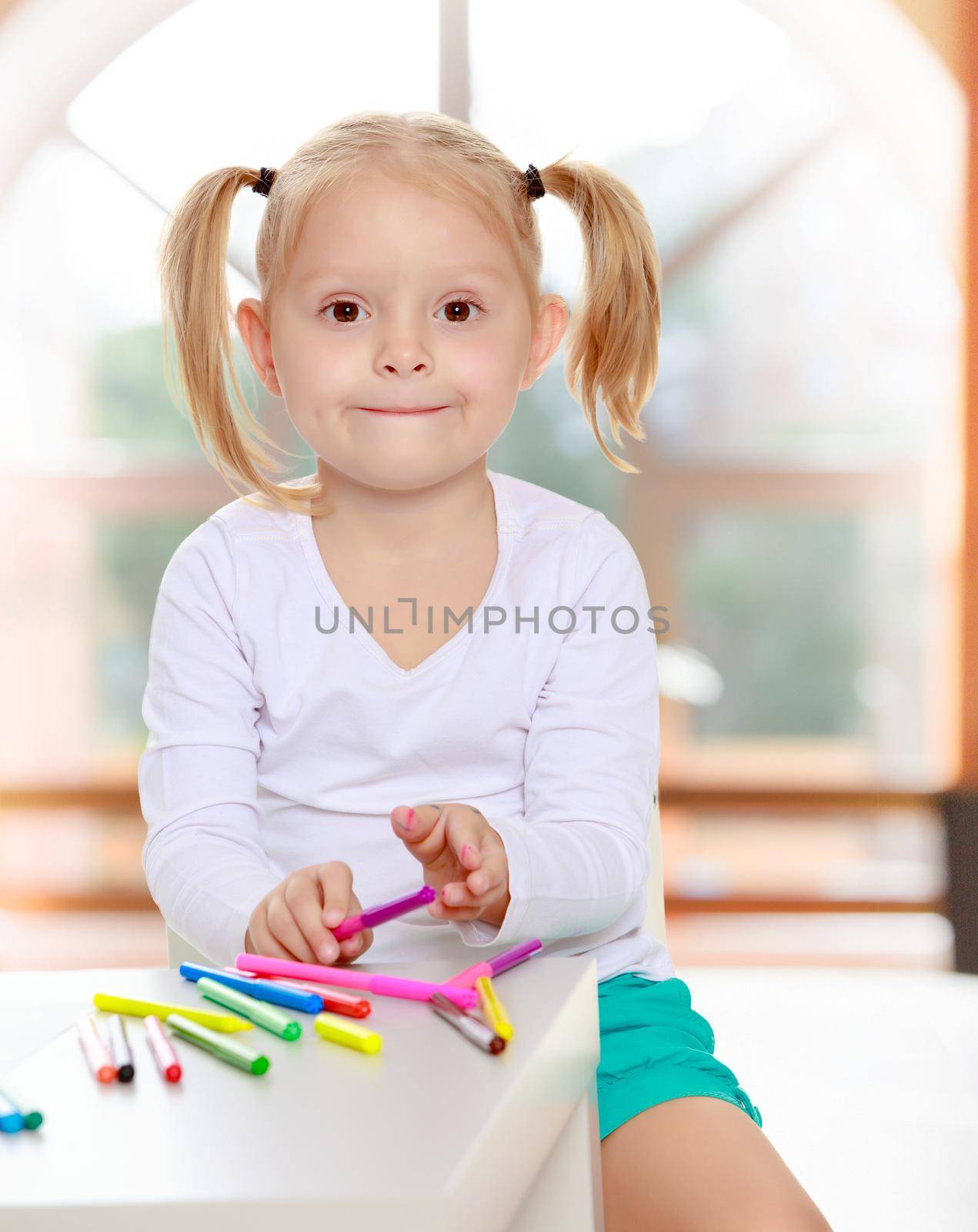 Pretty little blonde girl drawing with markers at the table.Girl holding in hands a pink marker.The concept of pre-school education of the child among their peers . in gaming room with a large arched window.