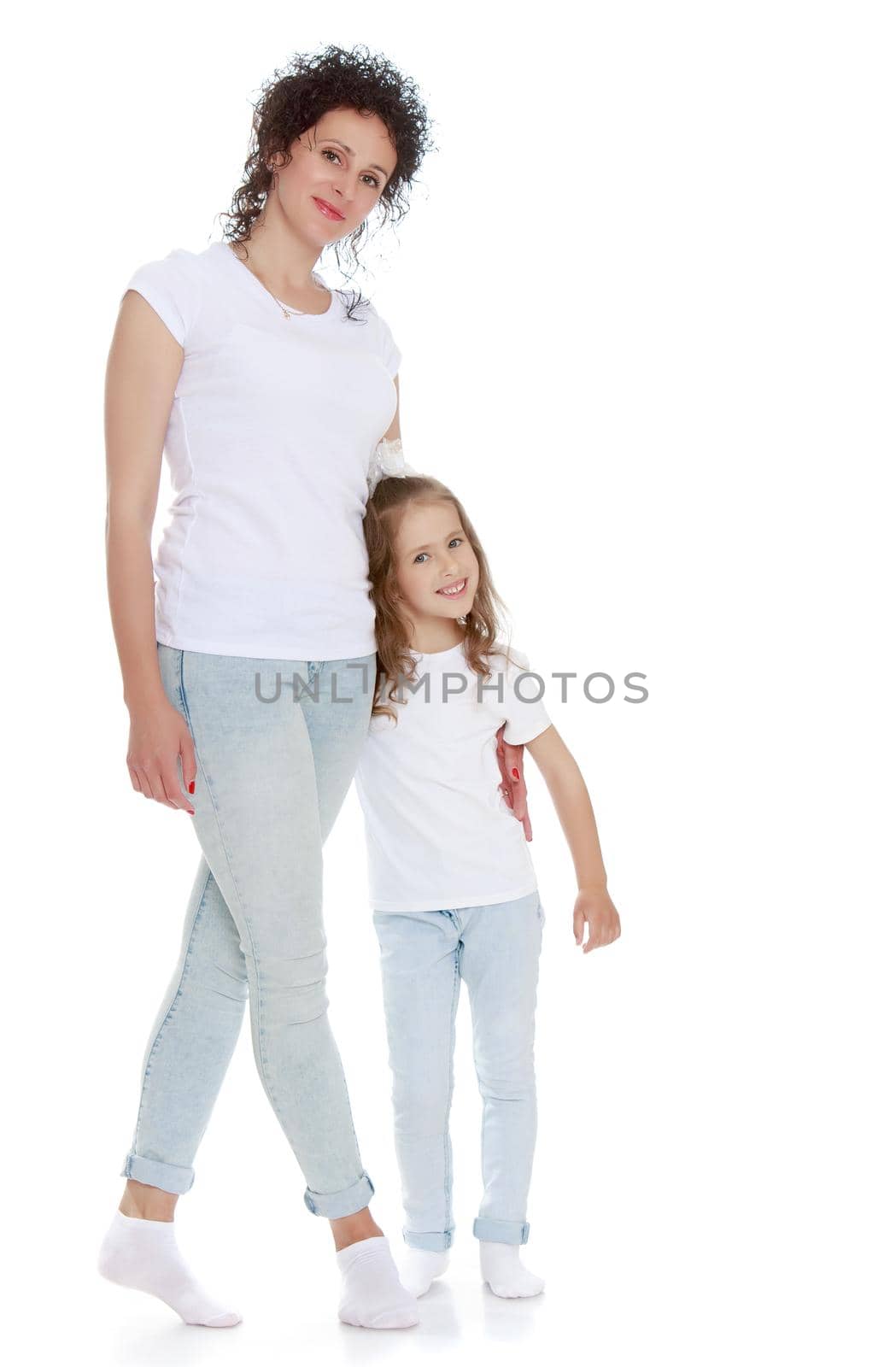 Beautiful, charming young mother with adorable little daughter. In the same jeans and white t-shirts without a pattern. Mother hugs daughter's waist - Isolated on white background
