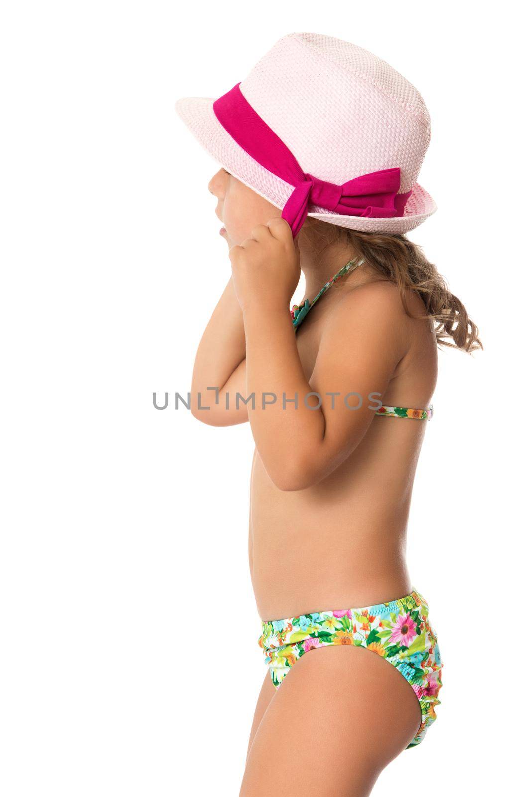 Gentle little girl in the pink hat and the swimsuit. The girl adjusts her hat with her hands. Turned sideways to the camera . Close-up - Isolated on white background