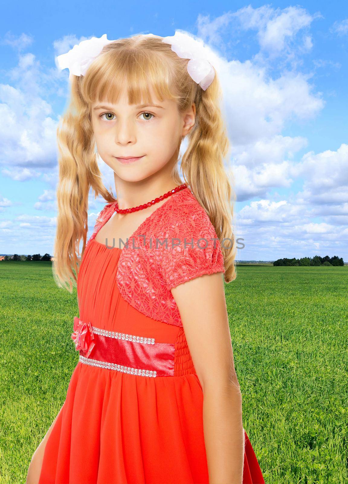 A very beautiful little girl with long, blonde ponytails on her head in a bright orange dress . close-up.On the background of bright green grass and blue sky with beautiful clouds.