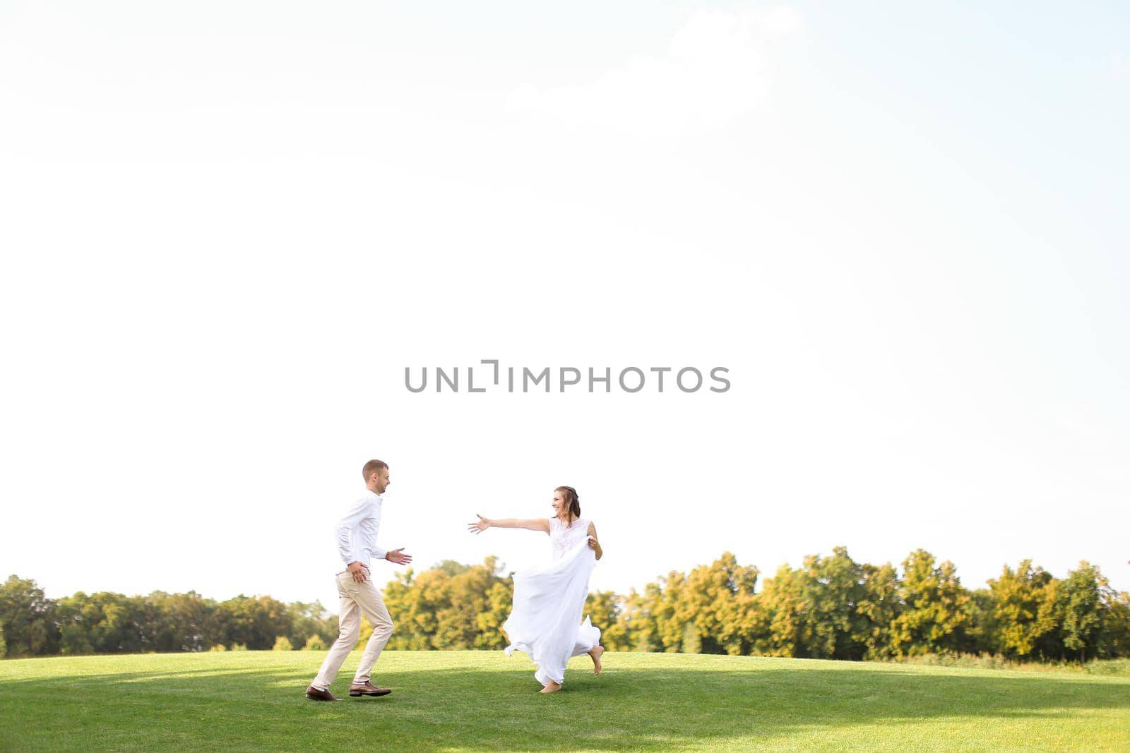 Groom and happy bride running and playing on grass. Concept of wedding photo session on open air and nature.
