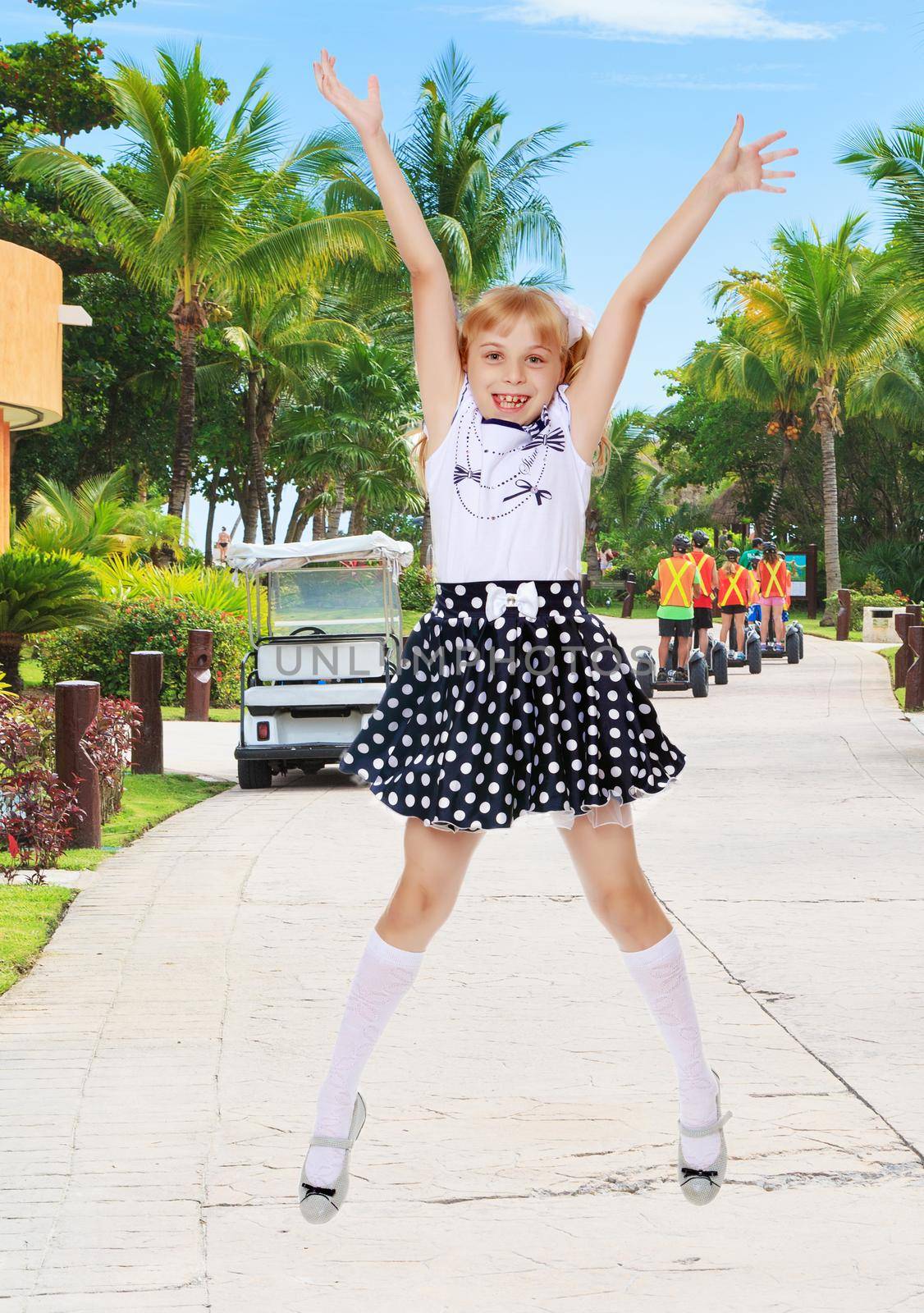 Cheerful little girl short skirt polka dot jumps with her arms and legs.On the background of the road, palm trees and blue sky with clouds.