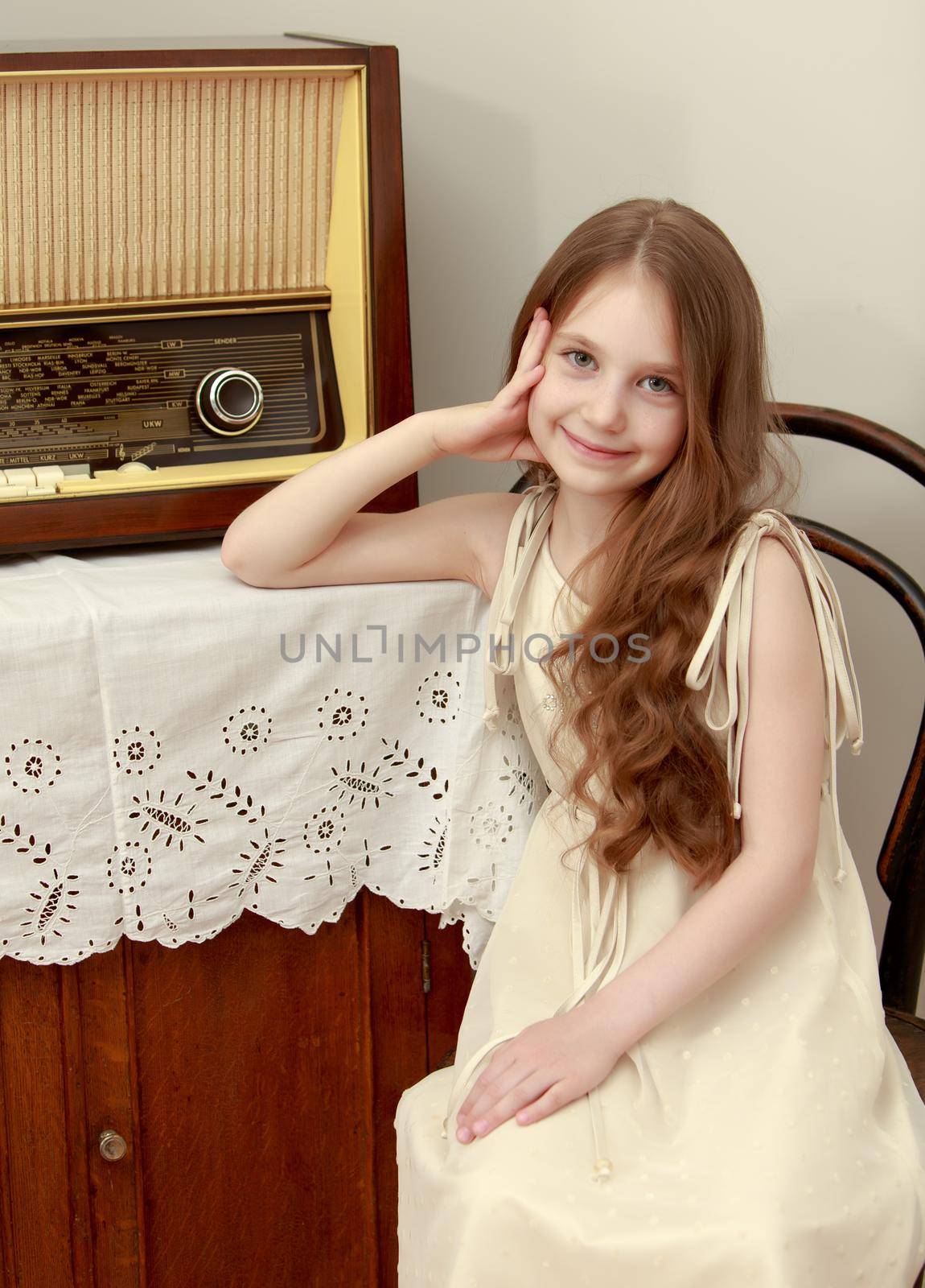 Gentle little girl with long brown hair to her waist . Girl sitting on an old Viennese chair near the old radio receiver