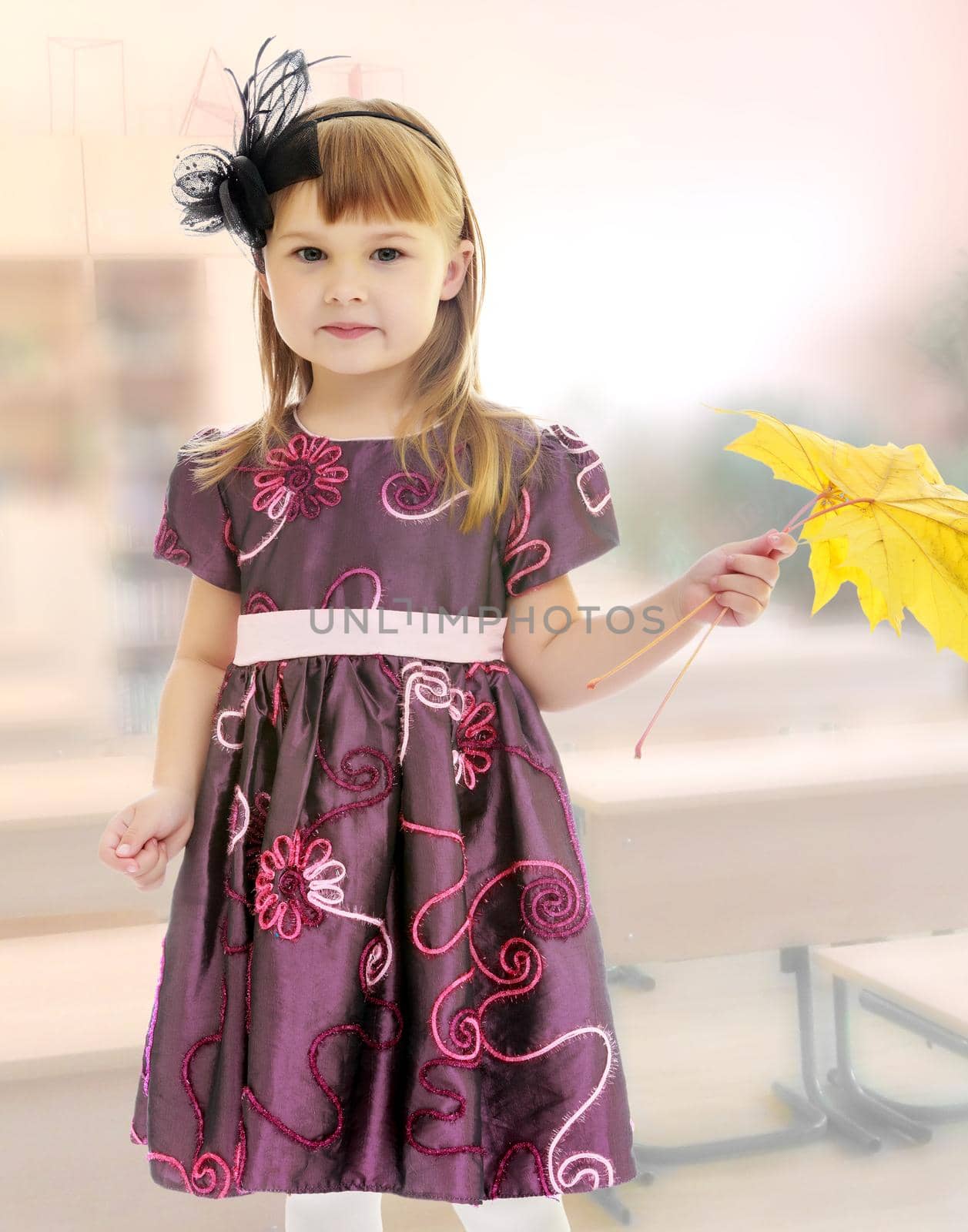 Adorable little girl holding a maple leaf.The concept of raising a child in the family, school and kindergarten. In the class where there are school desks.