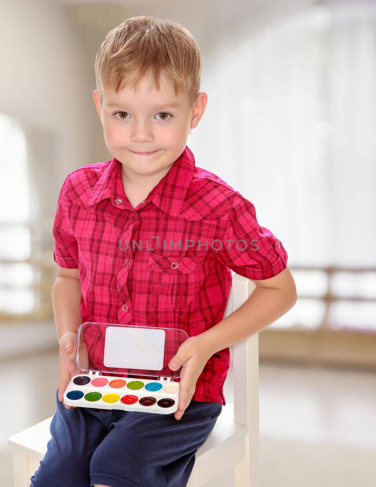 The concept of pre-school education of the child among their peers . in gaming room with a large arched window.Cute little boy in a red shirt holds his box of watercolors.