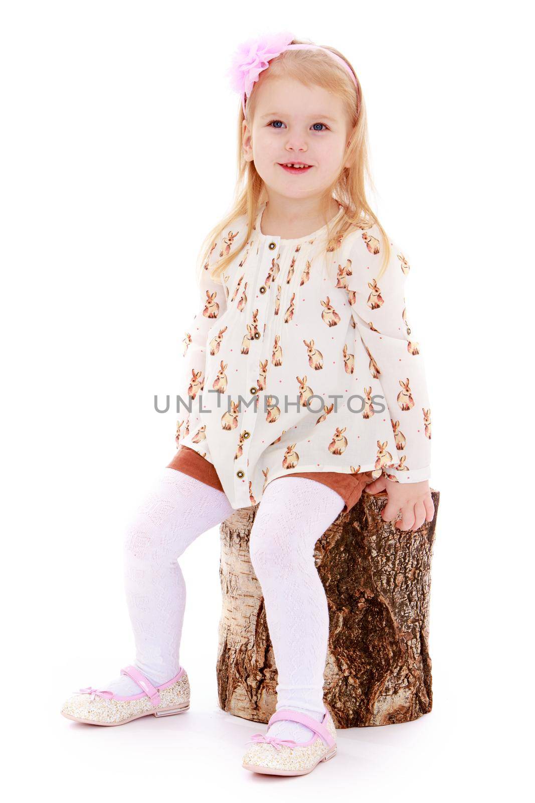 Cute little girl in shorts and white top thing with a big pink bow on her head. Girl sitting on a birch stump- Isolated on white background