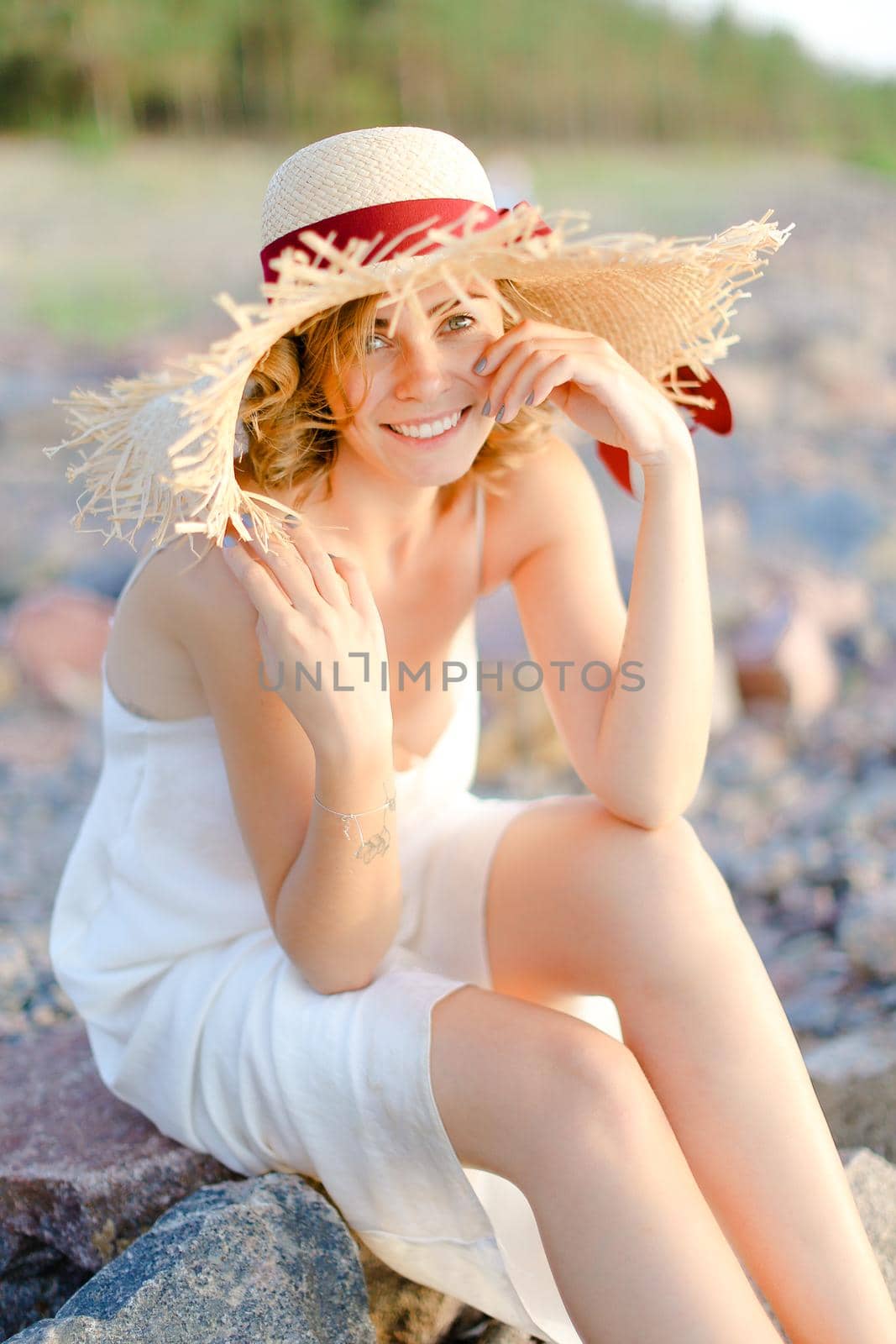 Caucasian nice girl in hat sitting on shingle beach. Concept of summer vacations and beauty.
