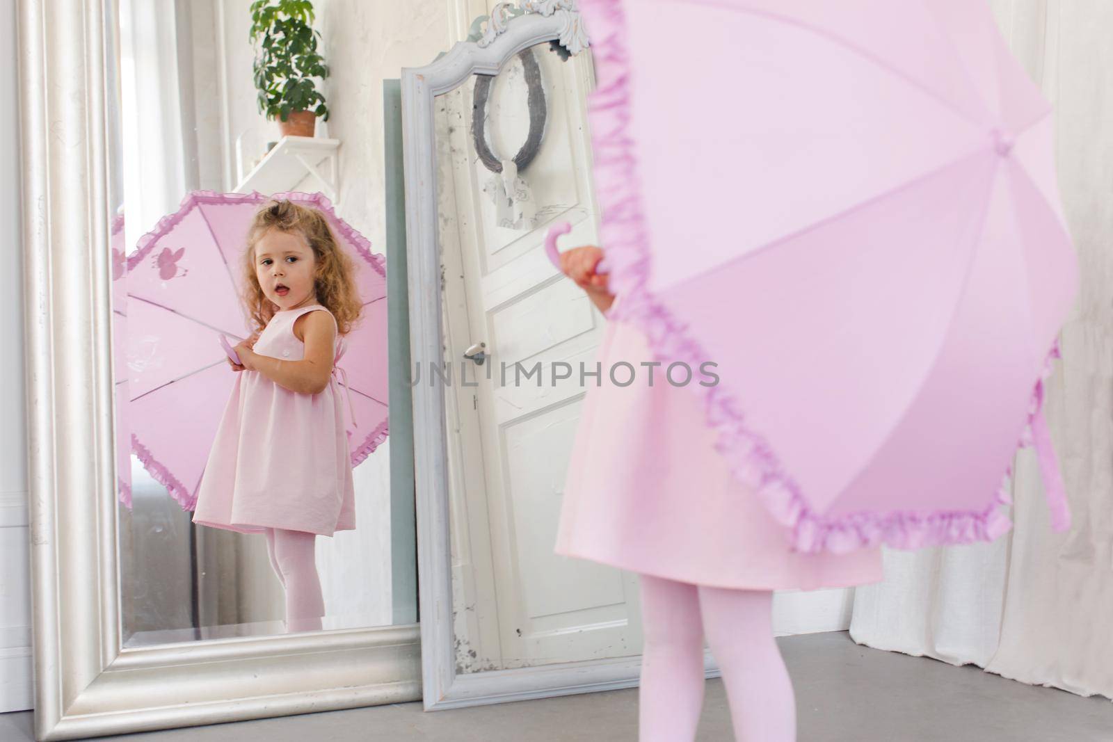 Adorable little child in pink dress and with umbrella looking in mirror while standing in cozy room