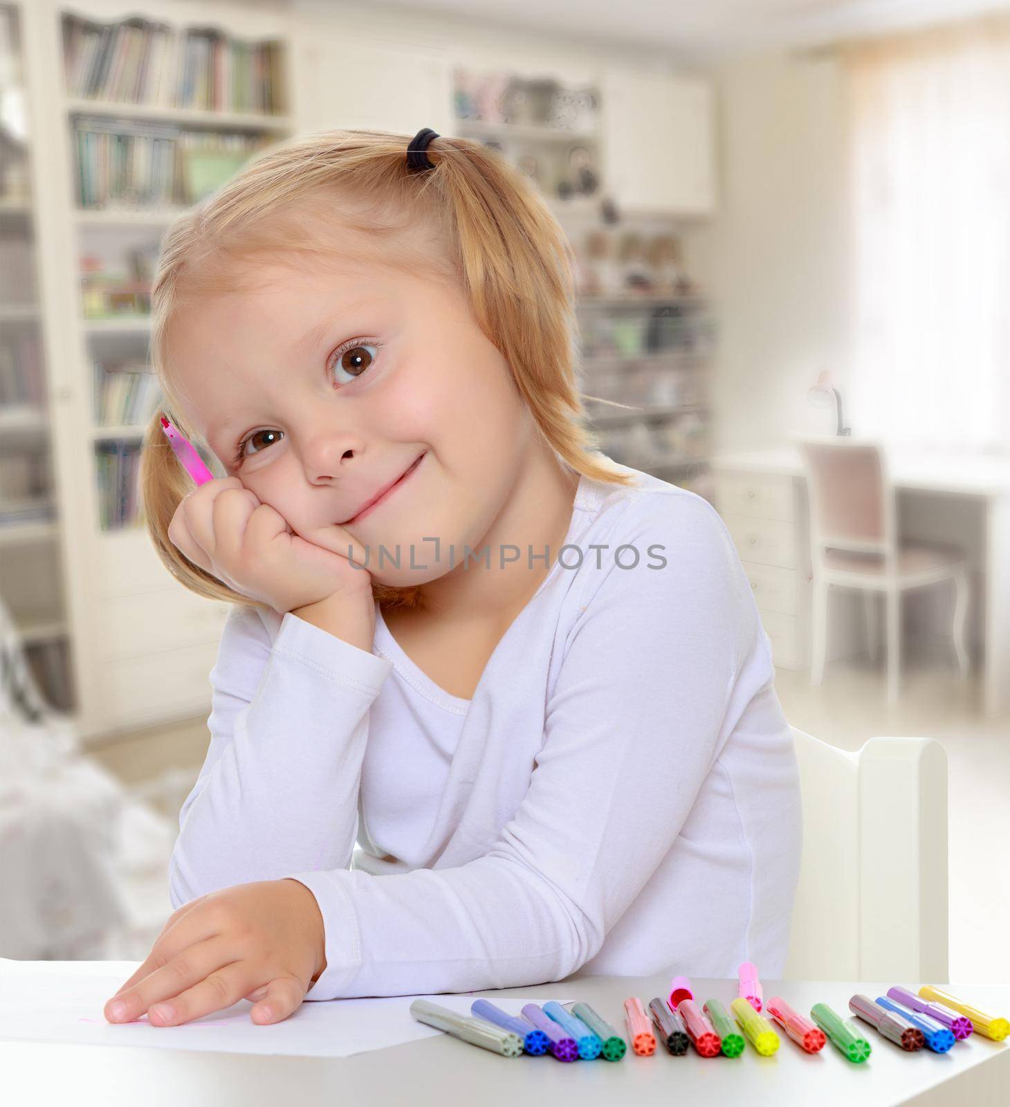 The concept of family happiness,and preschool education of the child , against a child's room with bookshelves.Pretty little blonde girl drawing with markers at the table.Girl put head on his hand