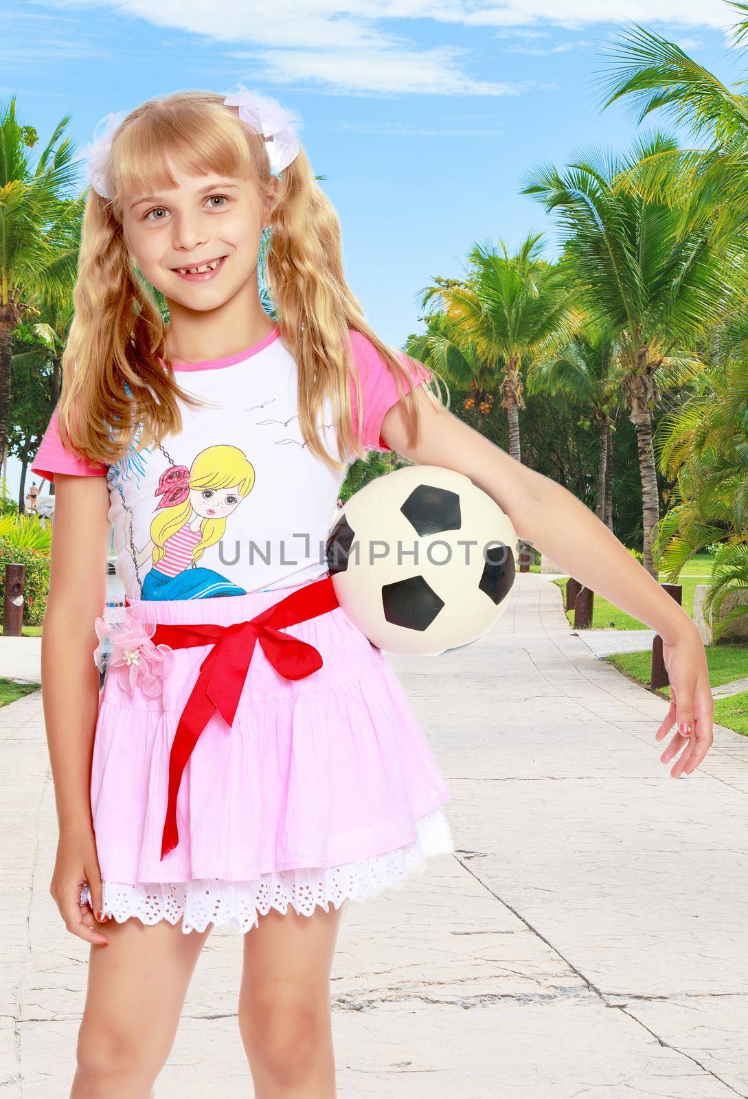 Close-up.Funny little blonde girl in a short pink skirt, holding a hand soccer ball.On the background of the road, palm trees and blue sky with clouds.