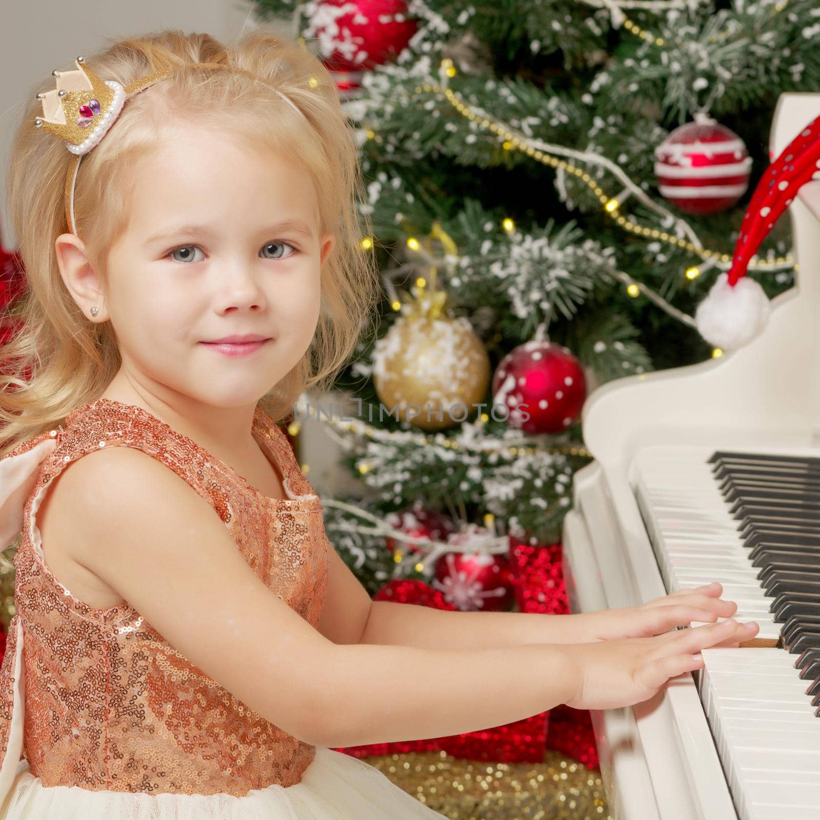 A cute little girl sitting near a white piano and a Christmas tree. The concept of the New Year, family holidays.