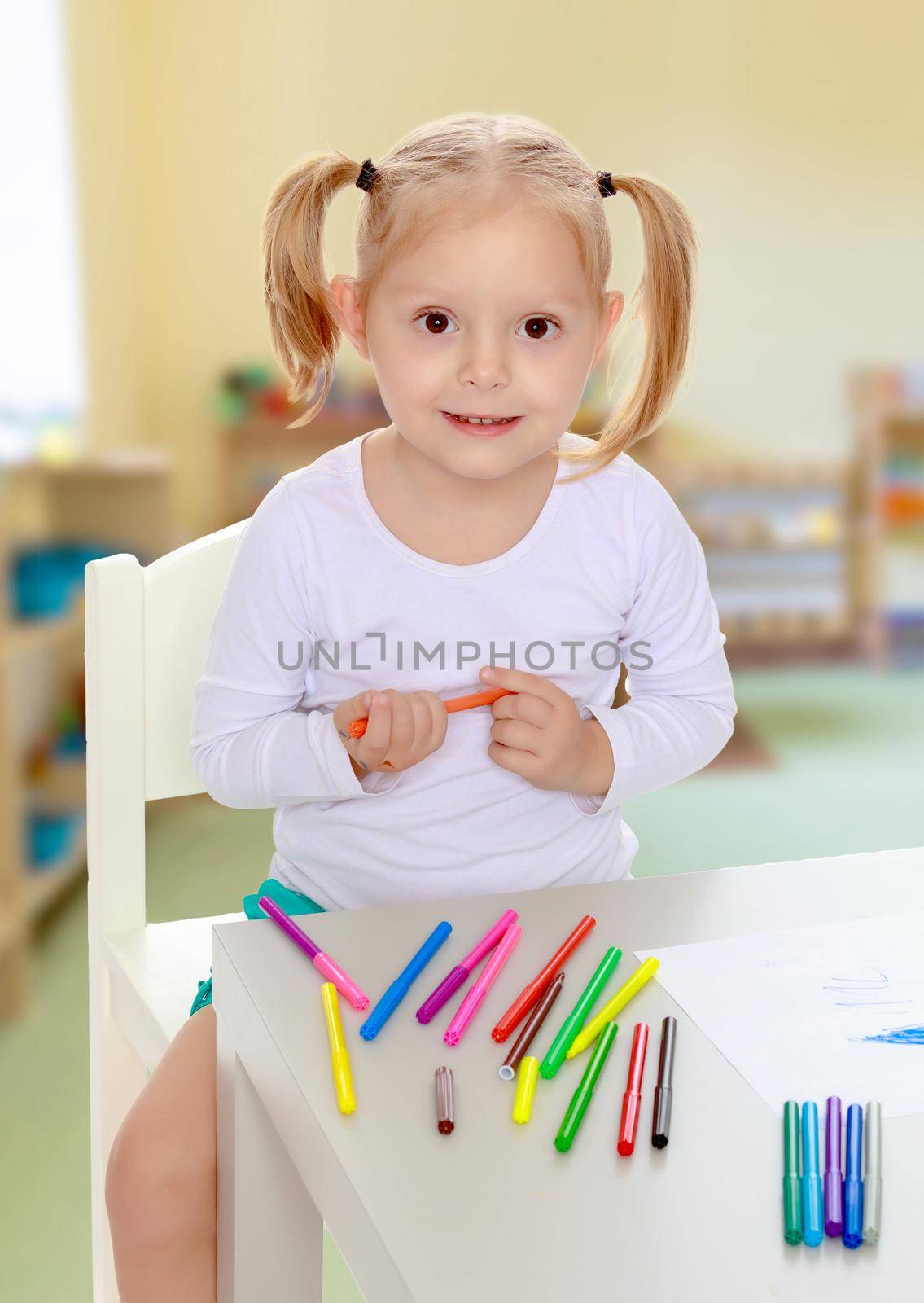 Pretty little blonde girl drawing with markers at the table.Girl holding in hands blue marker.The concept of pre-school education of the child among their peers . on the background of the playroom with shelves for toys. Montessori.
