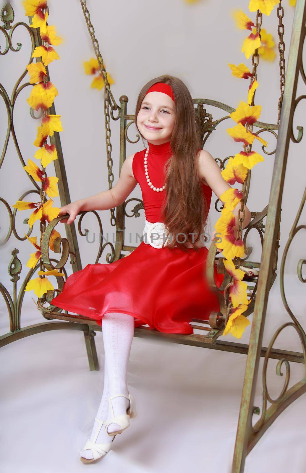Happy little girl with long brown hair to her waist . Girl in bright red dress. Girl swinging on a swing with his legs