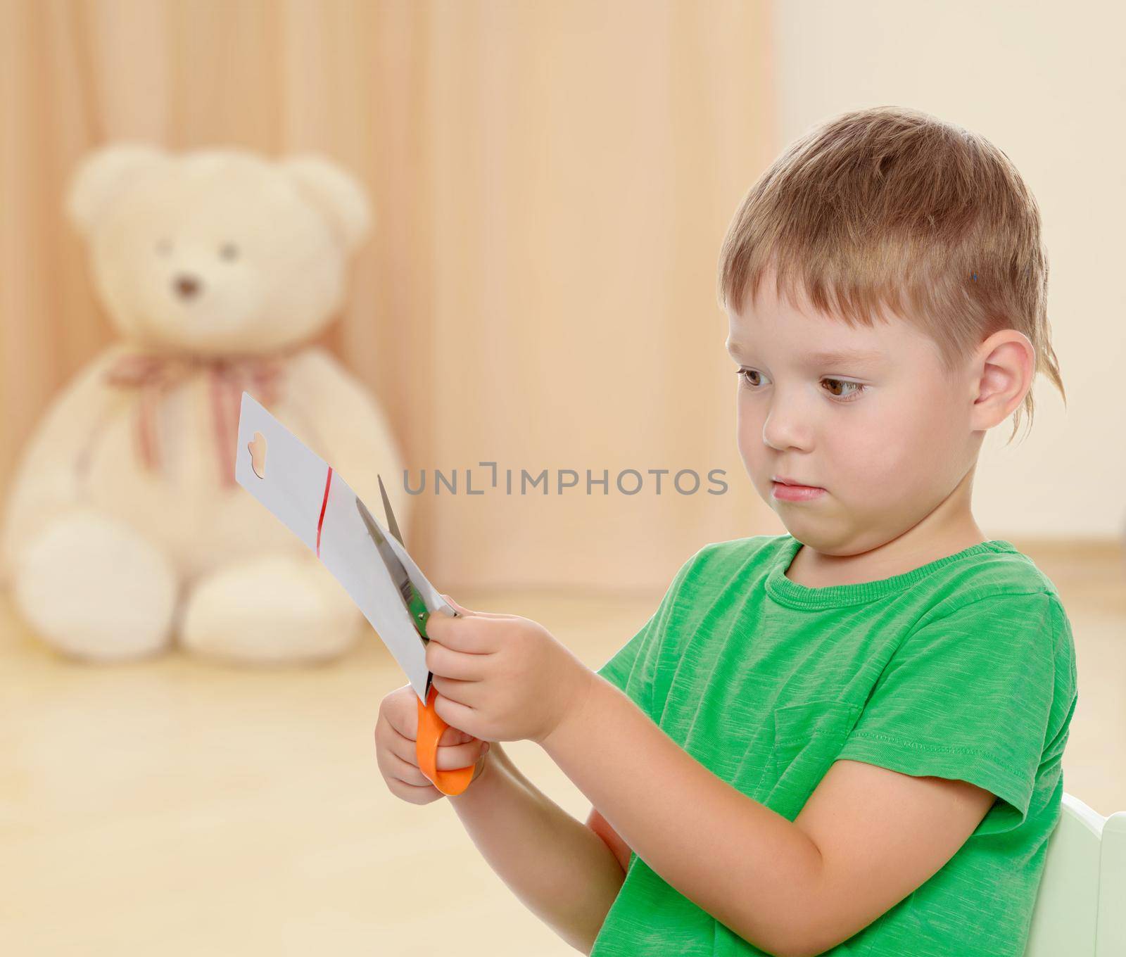 The concept of preschool development of the child ,against a child's room where in the background a Teddy bear.Cute little boy in the green shirt without a pattern , cut the cardboard with scissors.