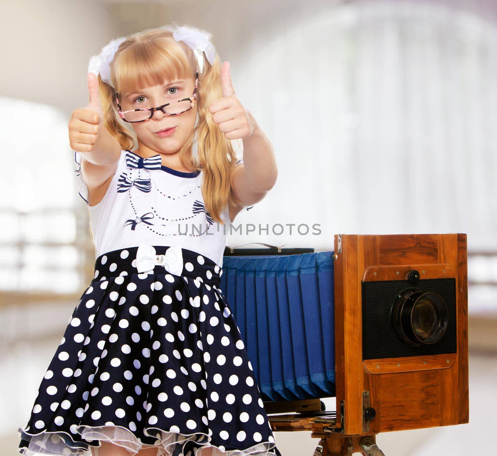Adorable little blond girl wearing glasses and fancy dress polka dot advertises the old wooden camera.In a room with a large semi-circular window.