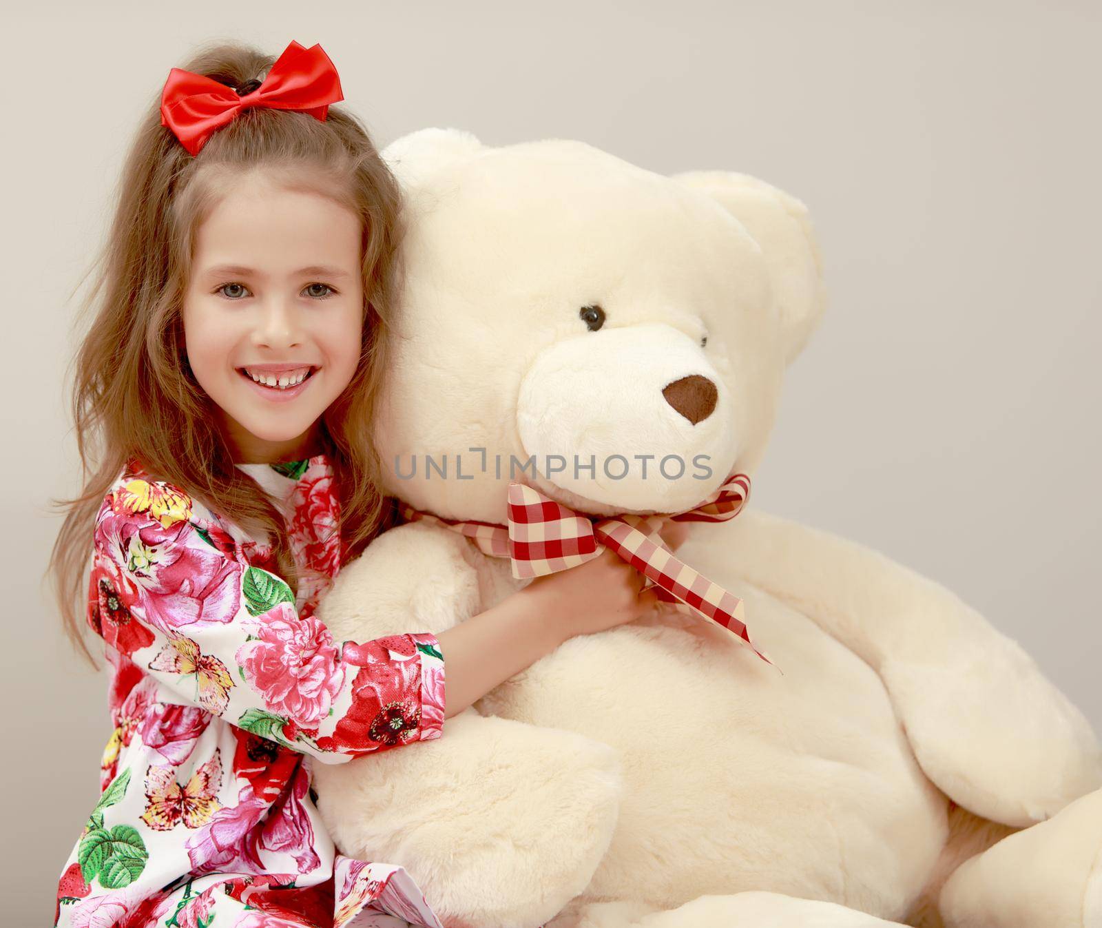 Happy little girl with a long ponytail down to his shoulders, and a red bow on her head, in a beautiful summer dress. Girl hugging a big Teddy bear. Close-up