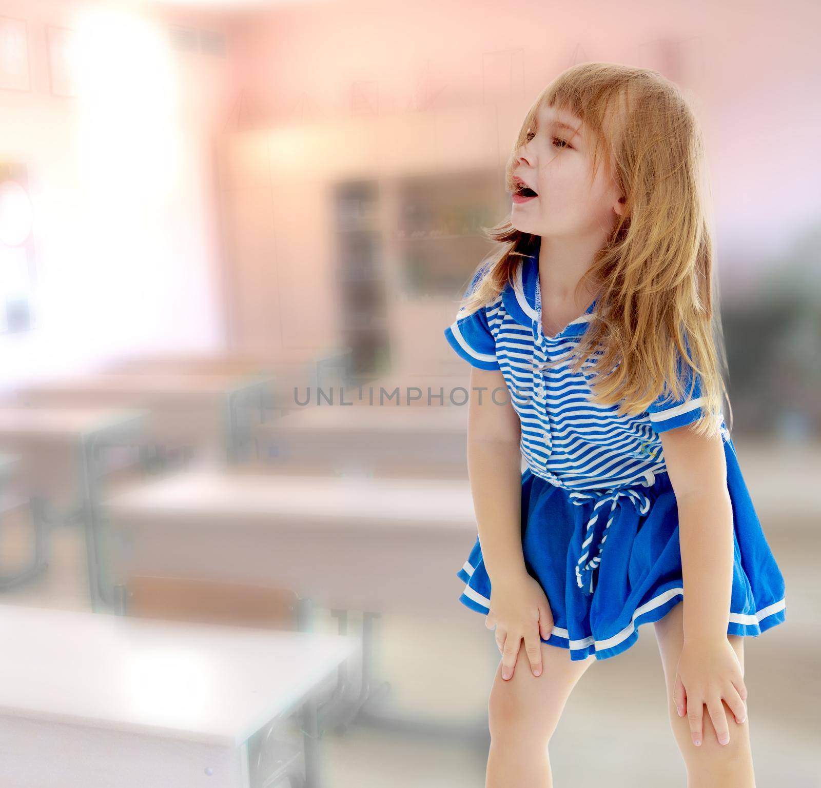 Cute little unkempt girl in a short blue dress. Girl looking to the side with his hands on his knees. In the class where there are school desks.