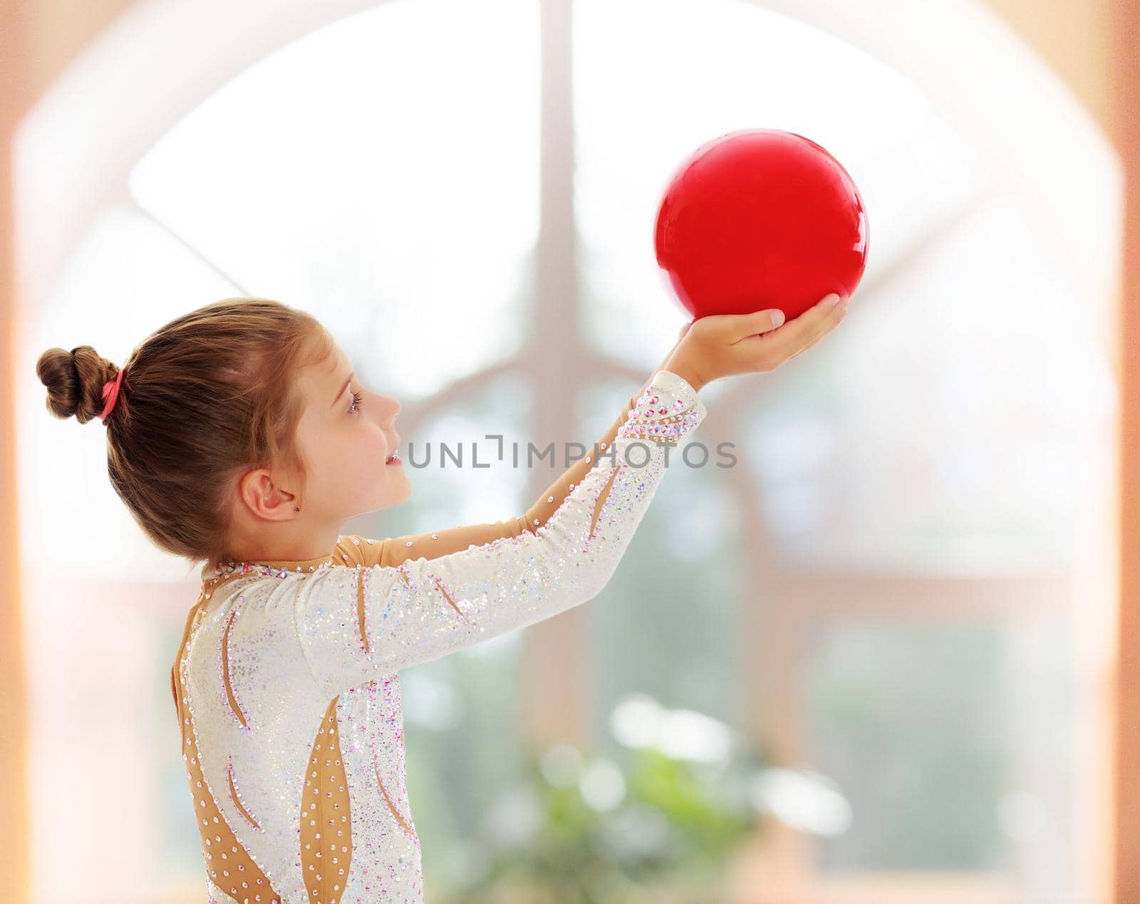On the background of the hall with large semi-circular window.Cute little girl gymnast turned sideways to the camera , holding in his outstretched hands a red ball.