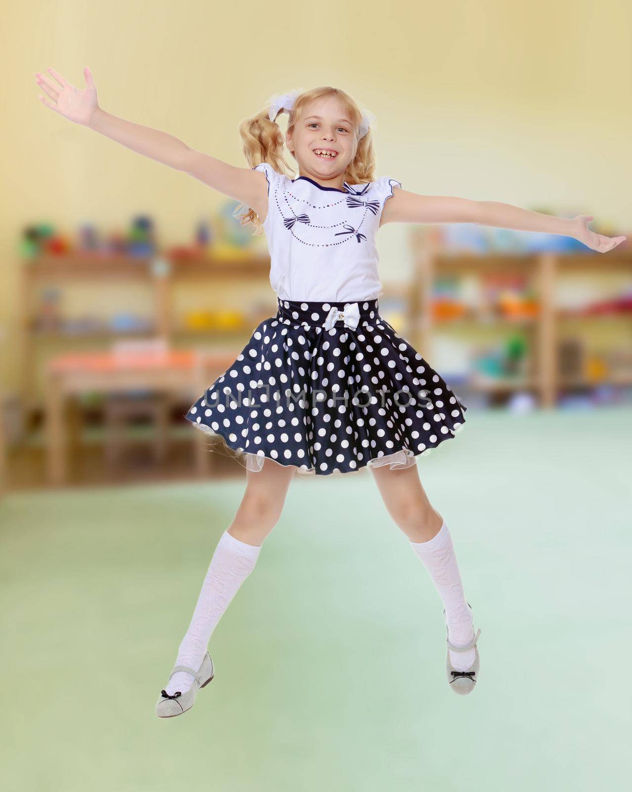 Happy little girl in the short polka-dot dress jumps , spread wide his arms and legs.In the background children's room where there are shelves with toys.