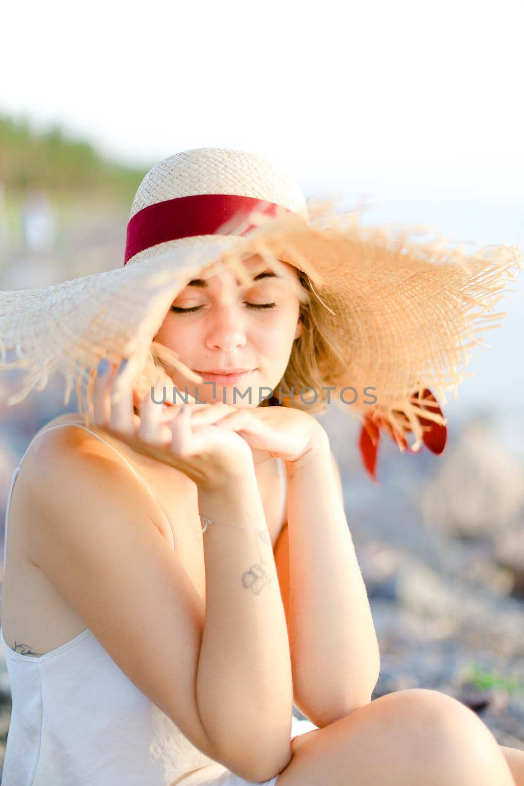 Portrait of beautiful woman wearing hat sitting on sand beach with closed eyes. Concept of fashion, beauty and summer vacations.