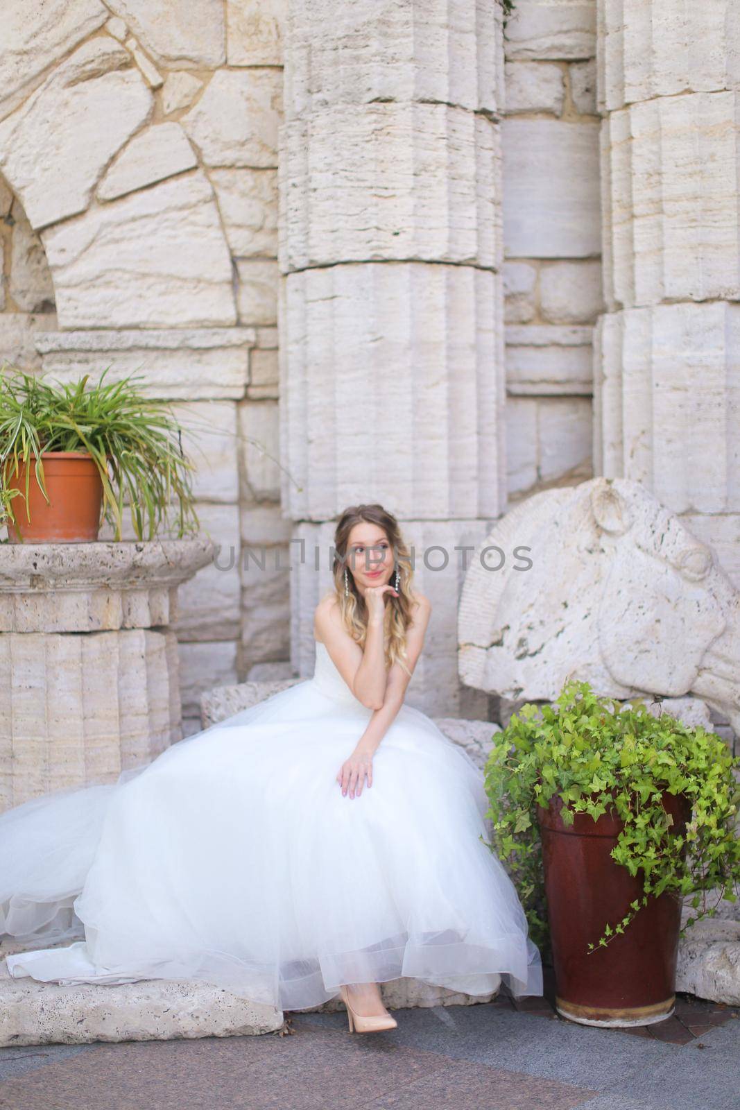 Caucasian european fiancee sitting near ancient columns and wearing white dress. Concept of bridal photo session and wedding.