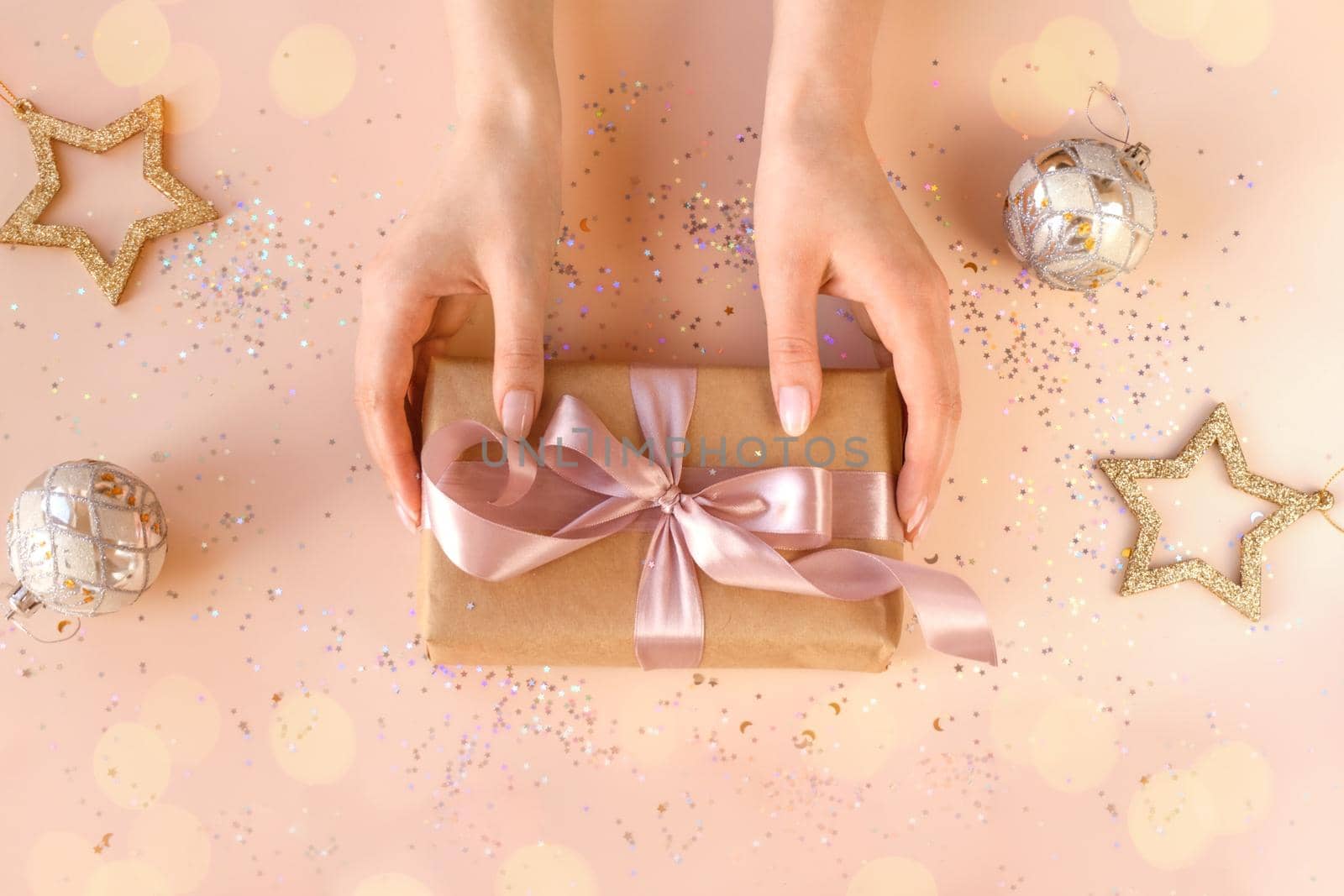 Female hands with natural manicure holding present in kraft paper with pink ribbon on soft beige background. Xmas composition. Flat lay. Happy holidays, New Year celebration and giving love concept.
