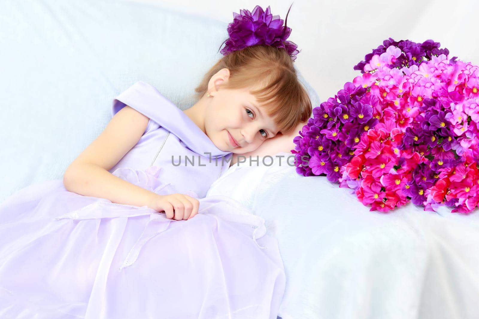 Fashionable little girl in a stylish beautiful dress.The girl sits on a luxurious white armchair near a large bouquet of flowers.
