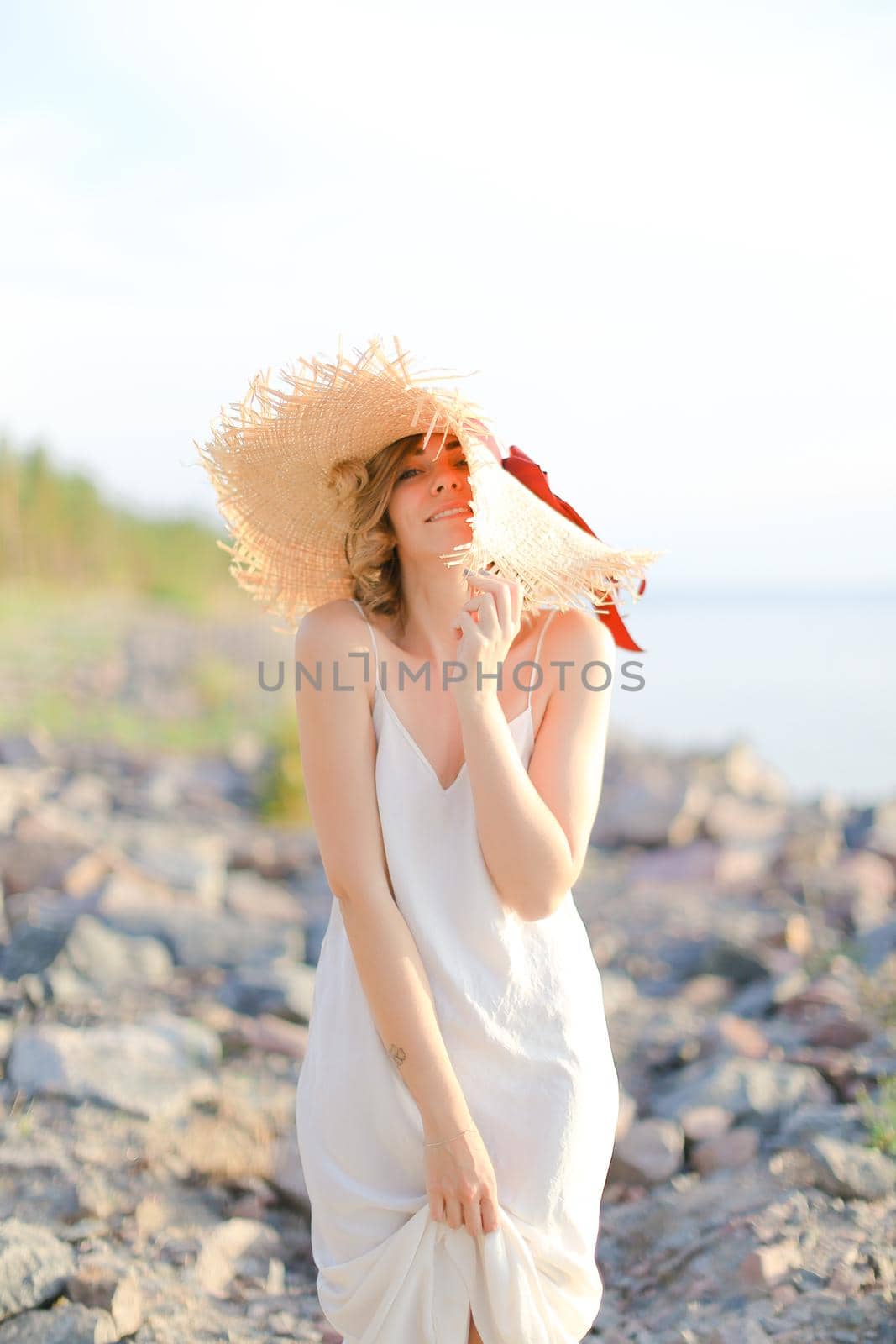 Young happy woman in hat with red ribbon standing on shingle beach. Concept of summer vacations and resort.
