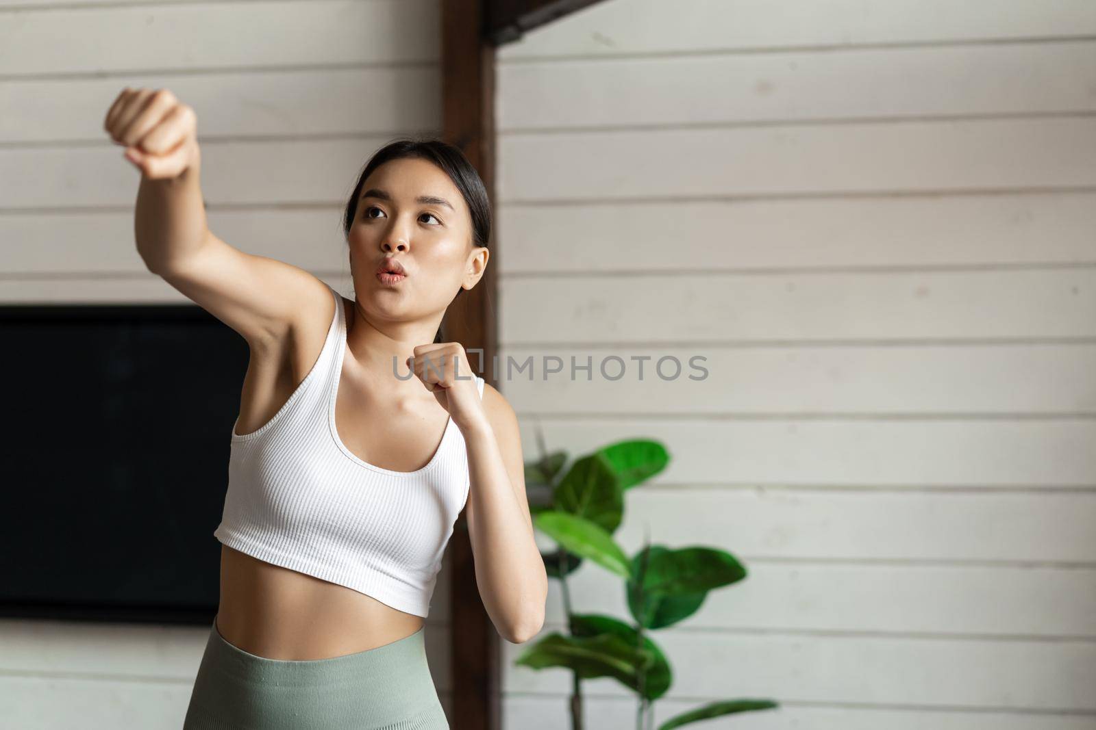 Fitness girl doing her workout at home. Asian athletic woman practice punches, wearing sport clothing.