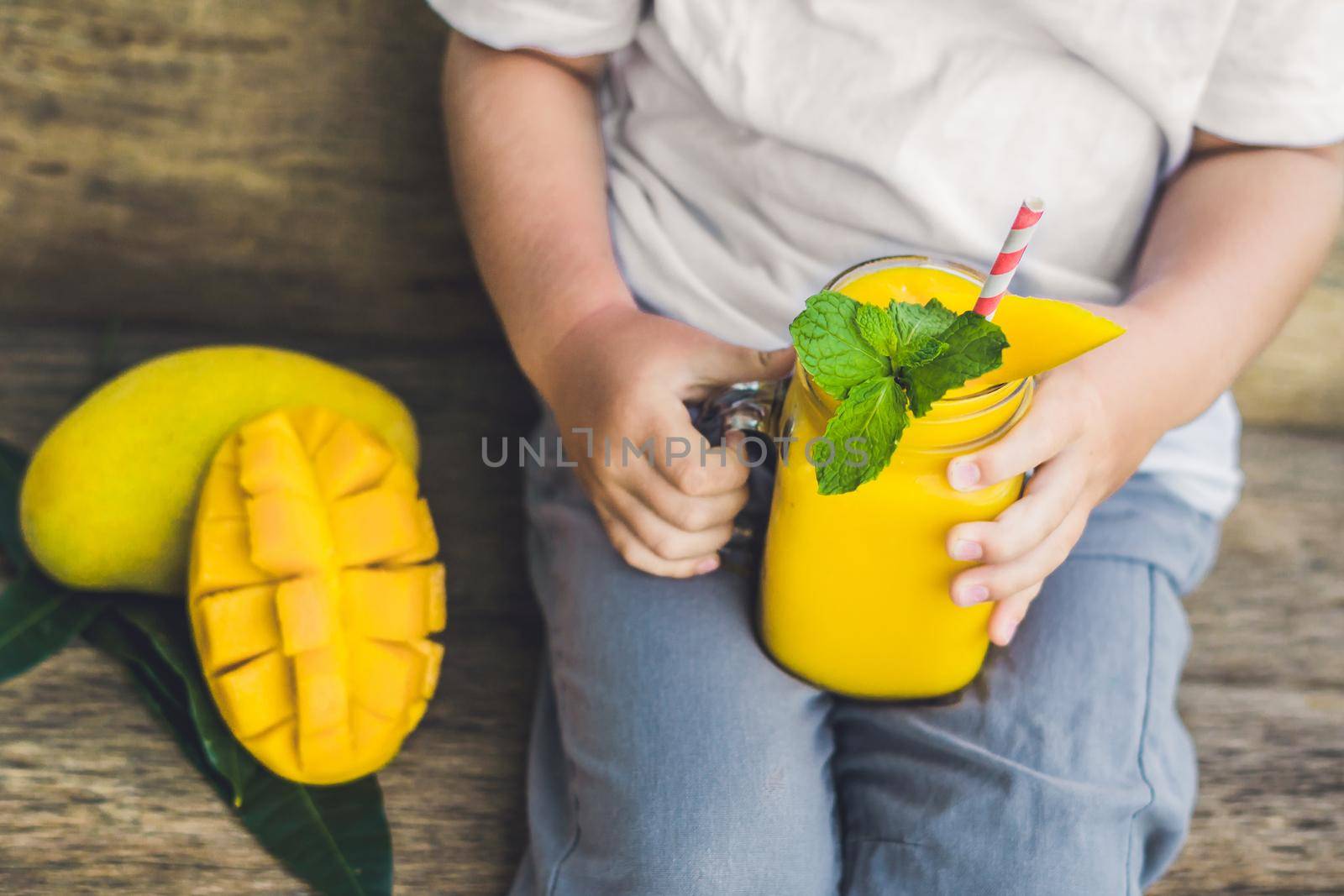 Boy drinking juicy smoothie from mango in glass mason jar with striped red straw on old wooden background. Healthy life concept, copy space.