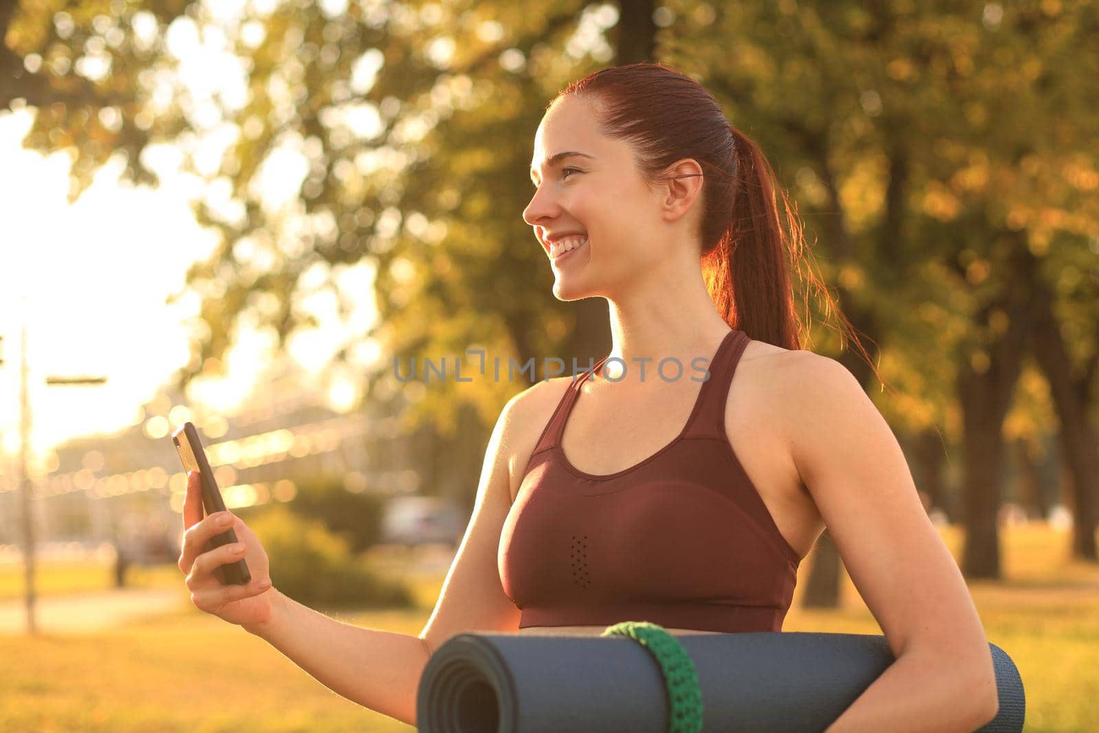 Cute smiling woman wearing tracksuit using smartphone and holding yoga mat at sunset in park