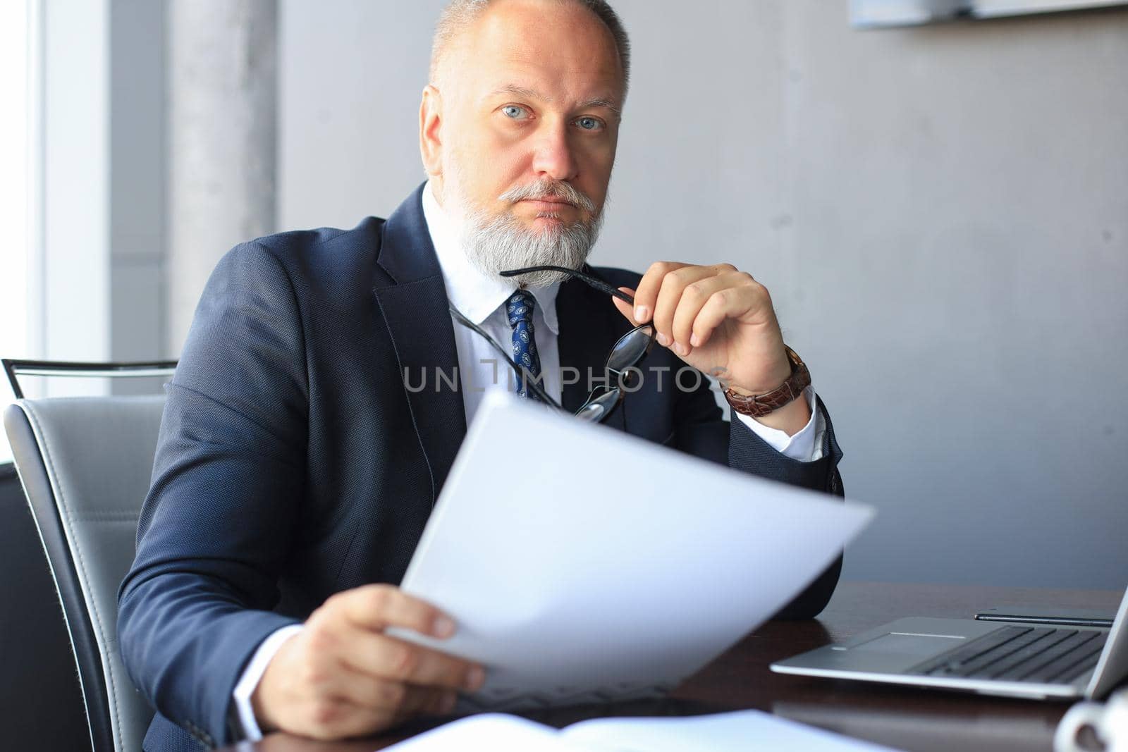 Mature business man in full suit holding his eyeglasses and looking at camera while sitting at the desk in office