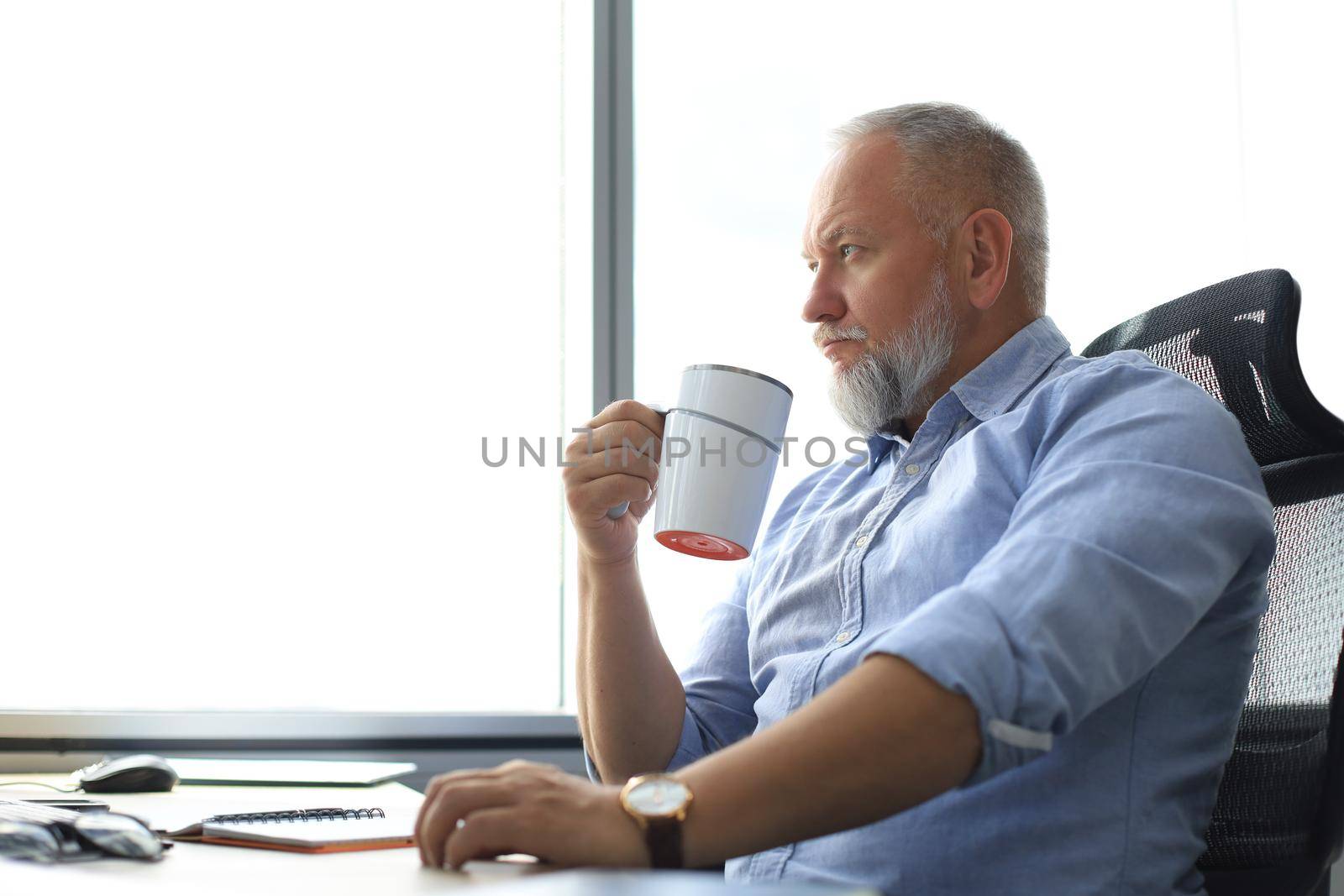 Focused mature businessman deep in thought while sitting at the desk with cup of coffee in his hand in modern office. by tsyhun
