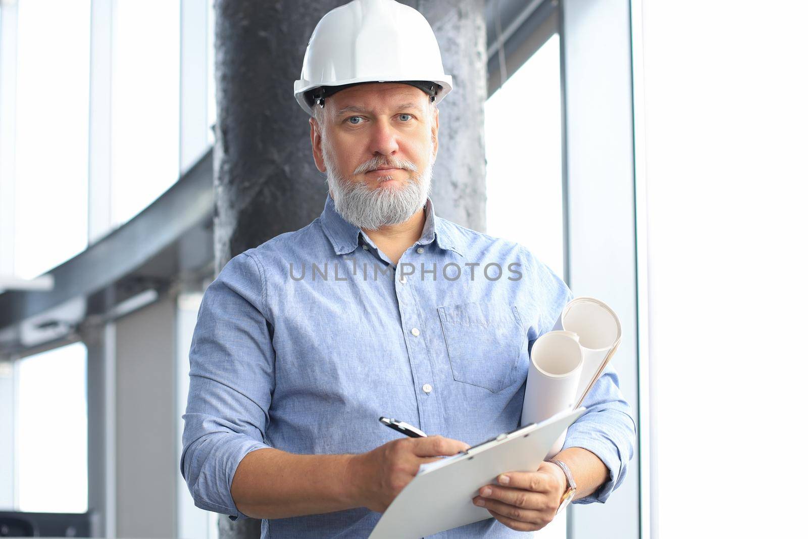 Confident mature business man in hardhat holding blueprint while standing indoors. by tsyhun