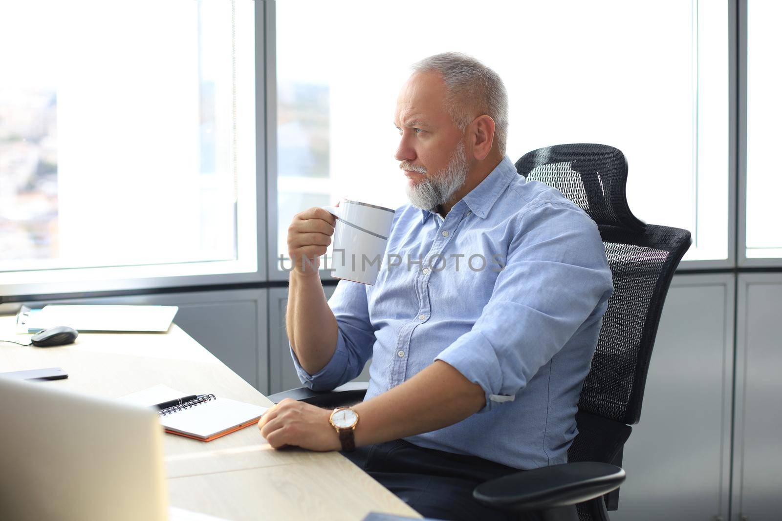 Focused mature businessman deep in thought while sitting at the desk with cup of coffee in his hand in modern office