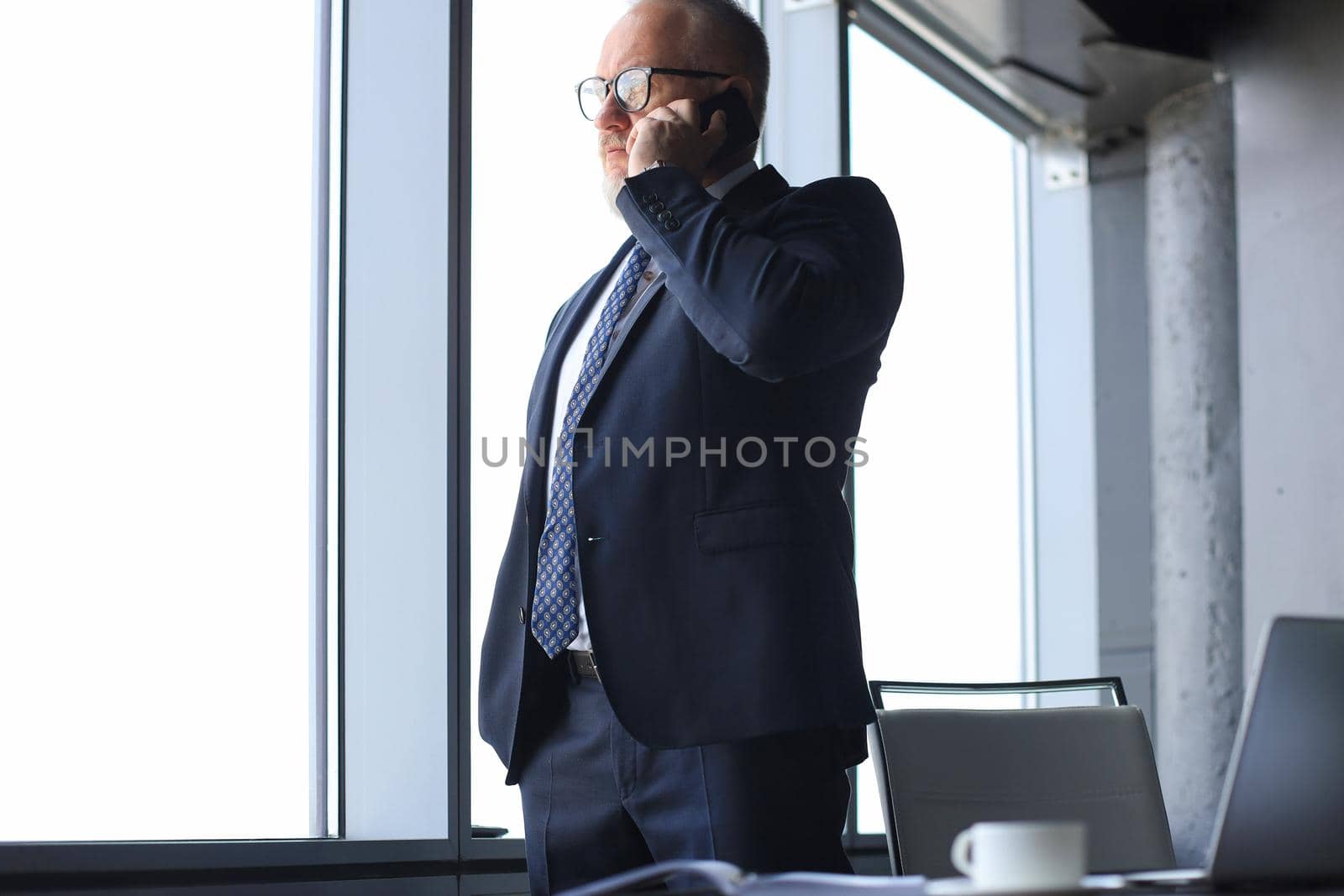 Mature businessman talking on a mobile phone standing by the window with view on city. by tsyhun